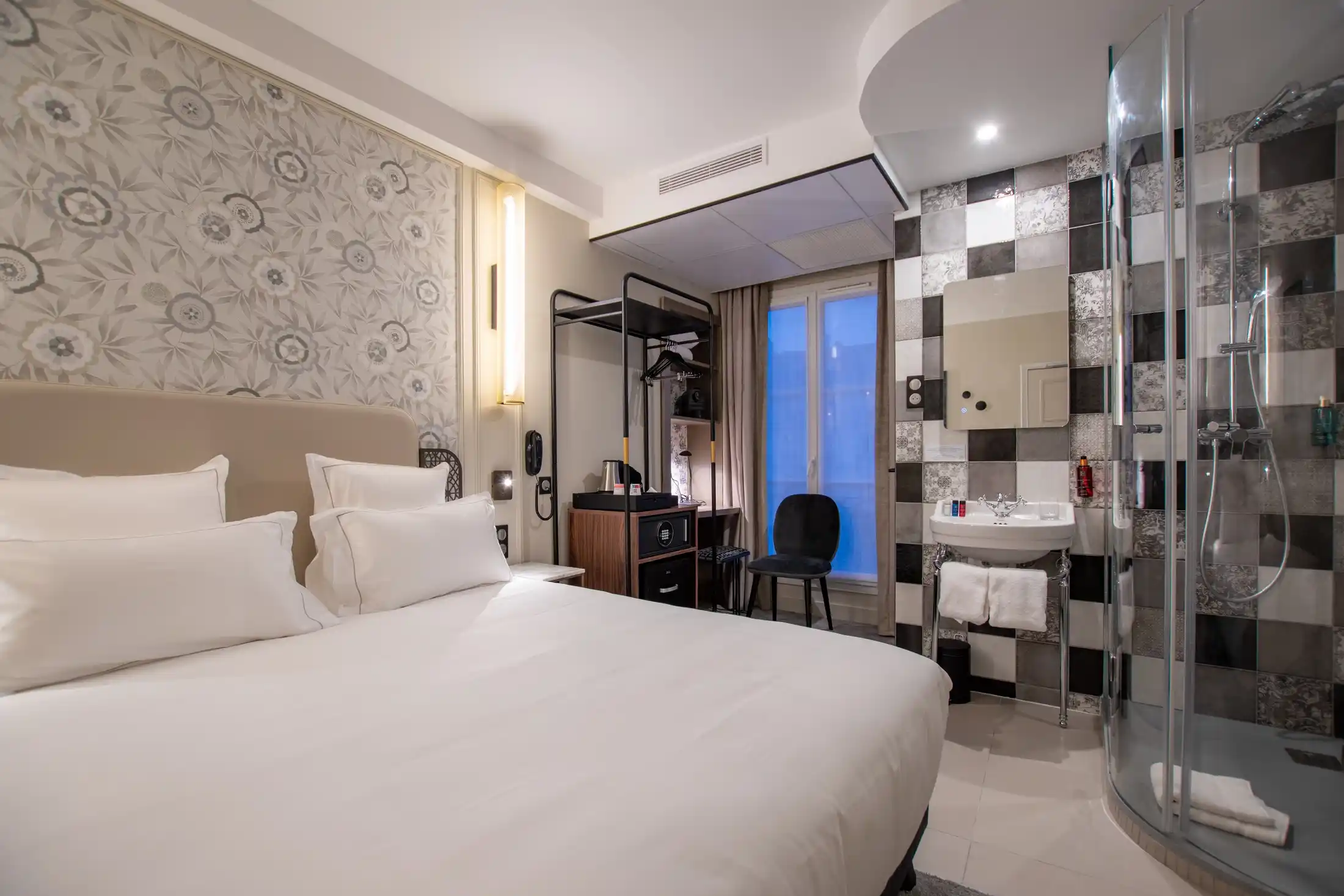 Quarto de hotel moderno com uma cama de casal grande com lençóis brancos, uma parede com padrão floral à esquerda, um móvel preto para roupas e uma escrivaninha sob uma janela, uma cadeira preta, uma pia com espelho em uma parede xadrez preta e branca e um box de vidro à direita.