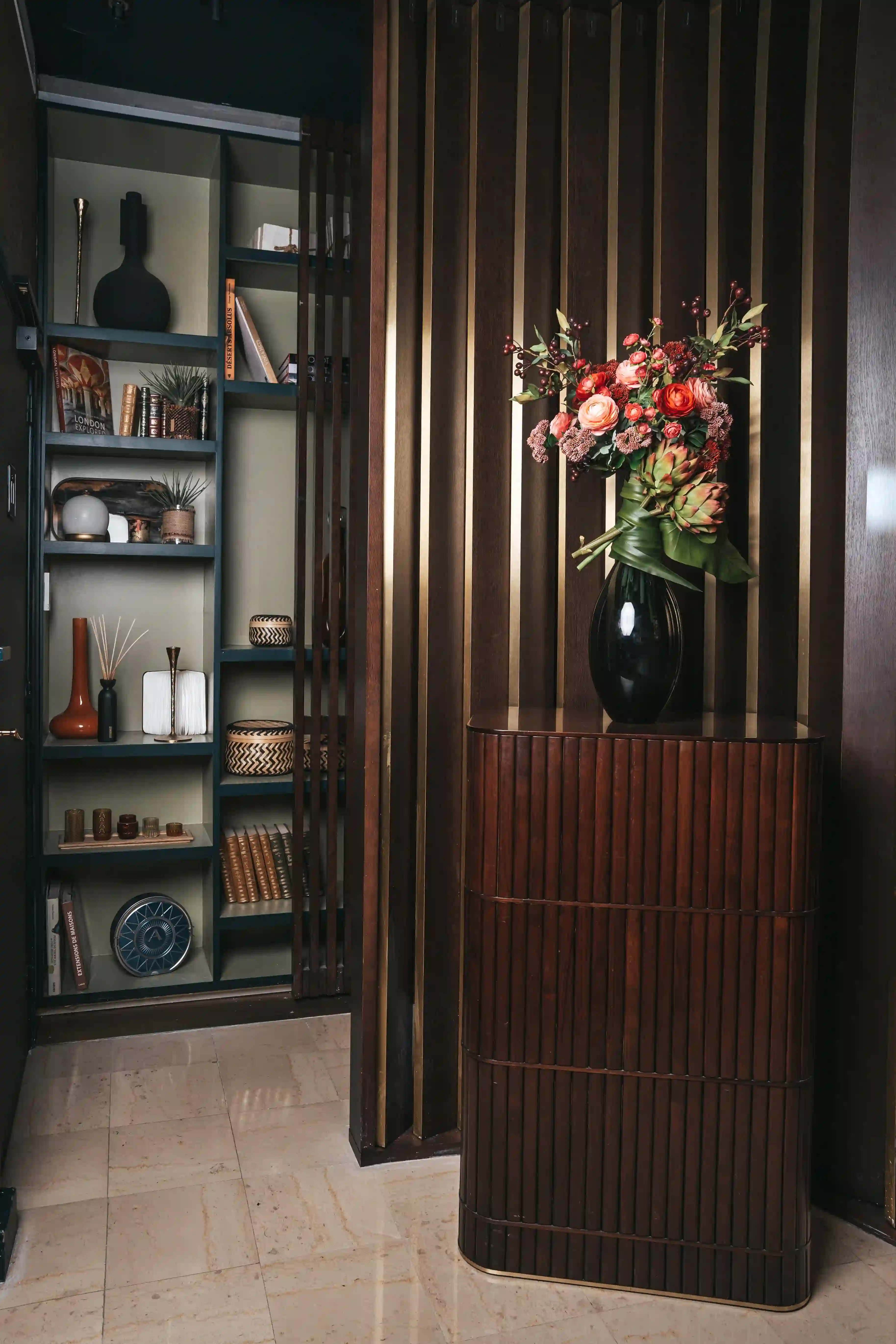 Interior elegante com um móvel de madeira escura com frisos verticais, sobreposto por um vaso preto com um bouquet colorido de flores frescas e folhagem verde, ao lado de uma estante de parede com livros, objetos decorativos e cestos de vime, tudo sobre um piso de mármore claro.