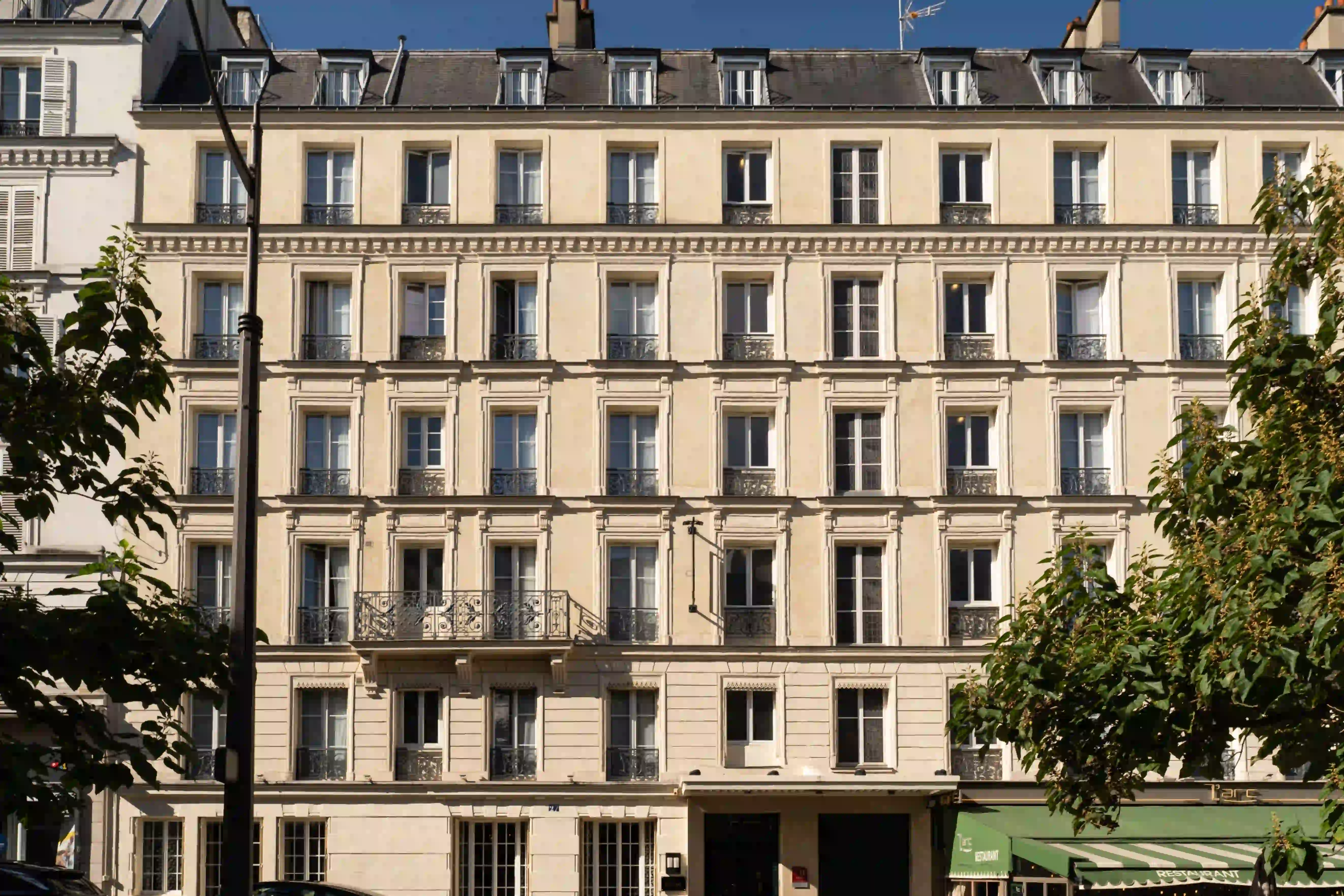 Fachada de um edifício residencial parisiense de estilo haussmanniano com vários andares de janelas com pequenas varandas de ferro forjado, sob um céu azul, emoldurada por árvores verdes.