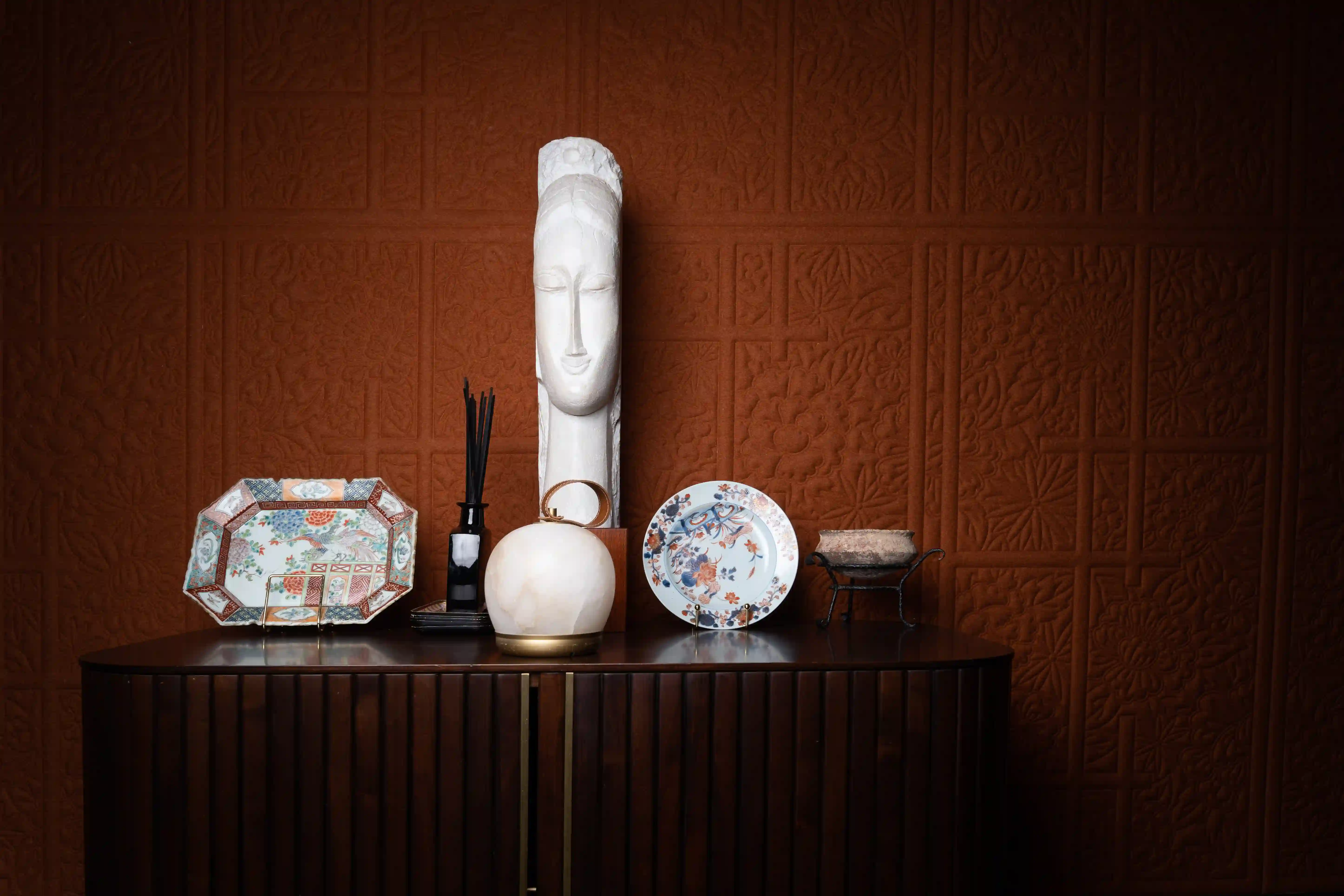 Um aparador escuro com objetos decorativos, incluindo uma escultura branca alongada de um rosto, dois pratos decorativos em suportes, um difusor com varetas pretas e um candeeiro branco redondo, em frente a uma parede castanha texturada com motivos florais.