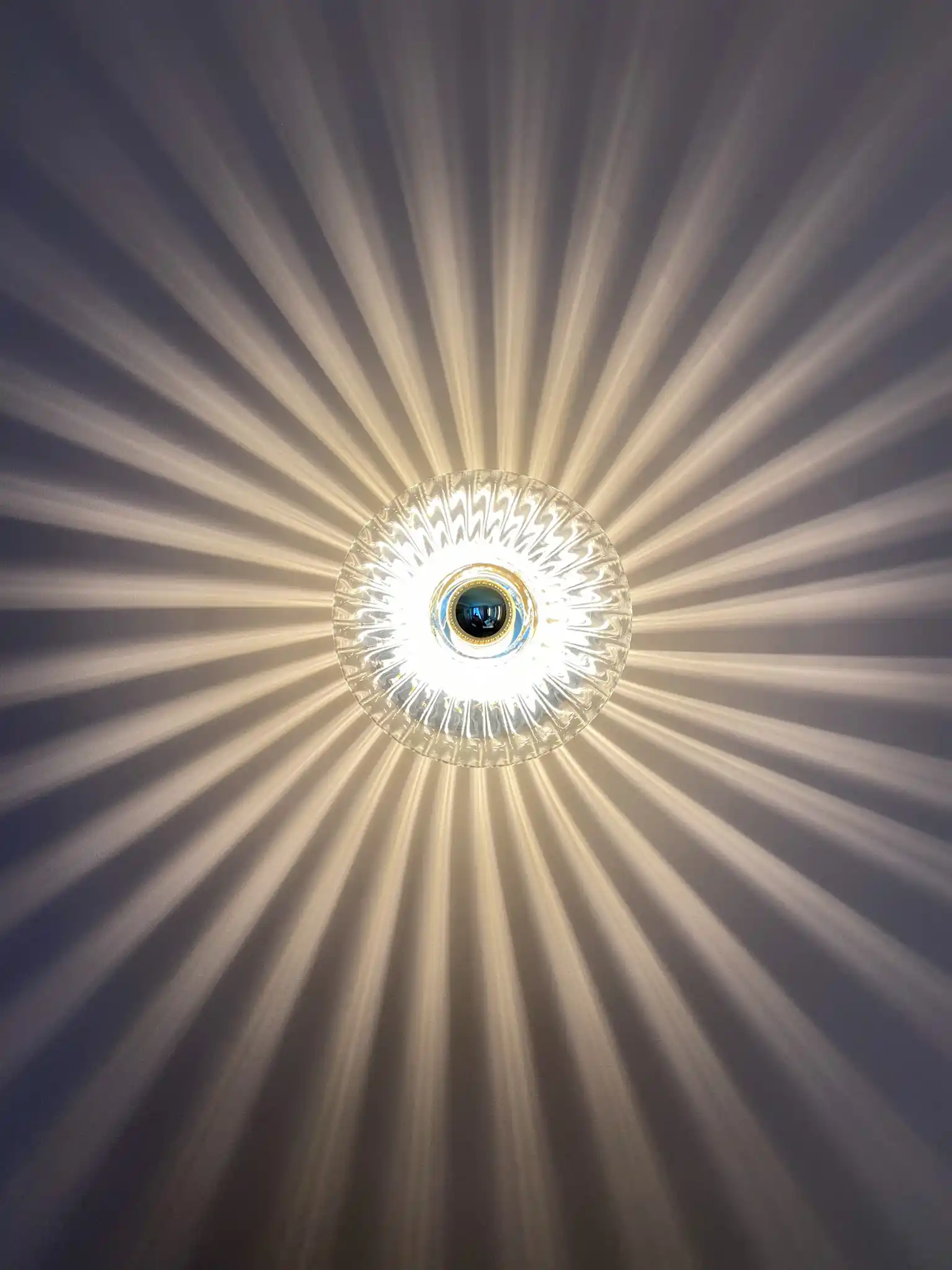 Luminária de teto em cristal transparente que projeta um padrão radiante de luz branca em uma parede cinza.