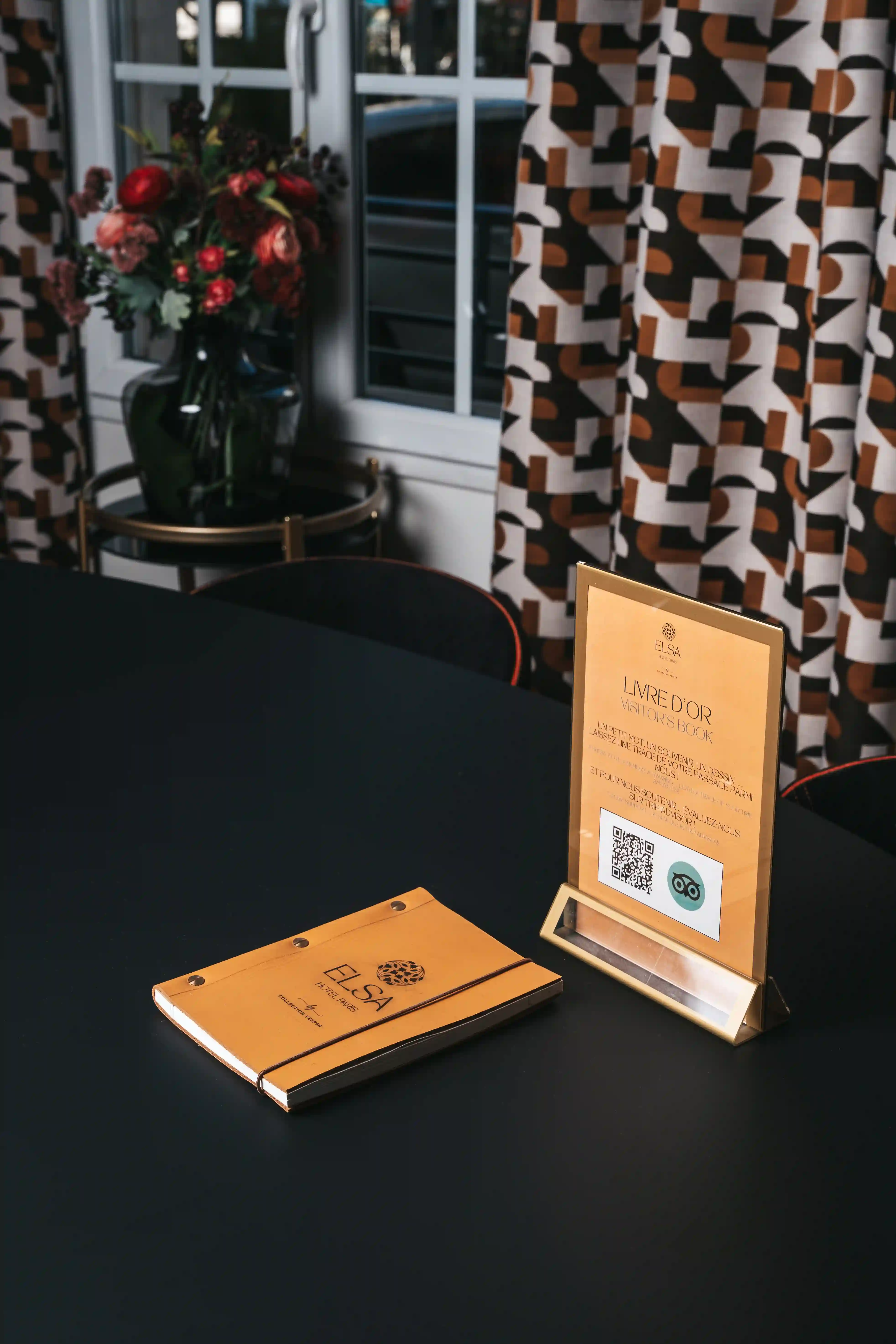 Livro de visitas laranja e cavalete com instruções para deixar uma mensagem e digitalizar um código QR sobre uma mesa preta num interior com um vaso de flores e cortinas com padrões geométricos.