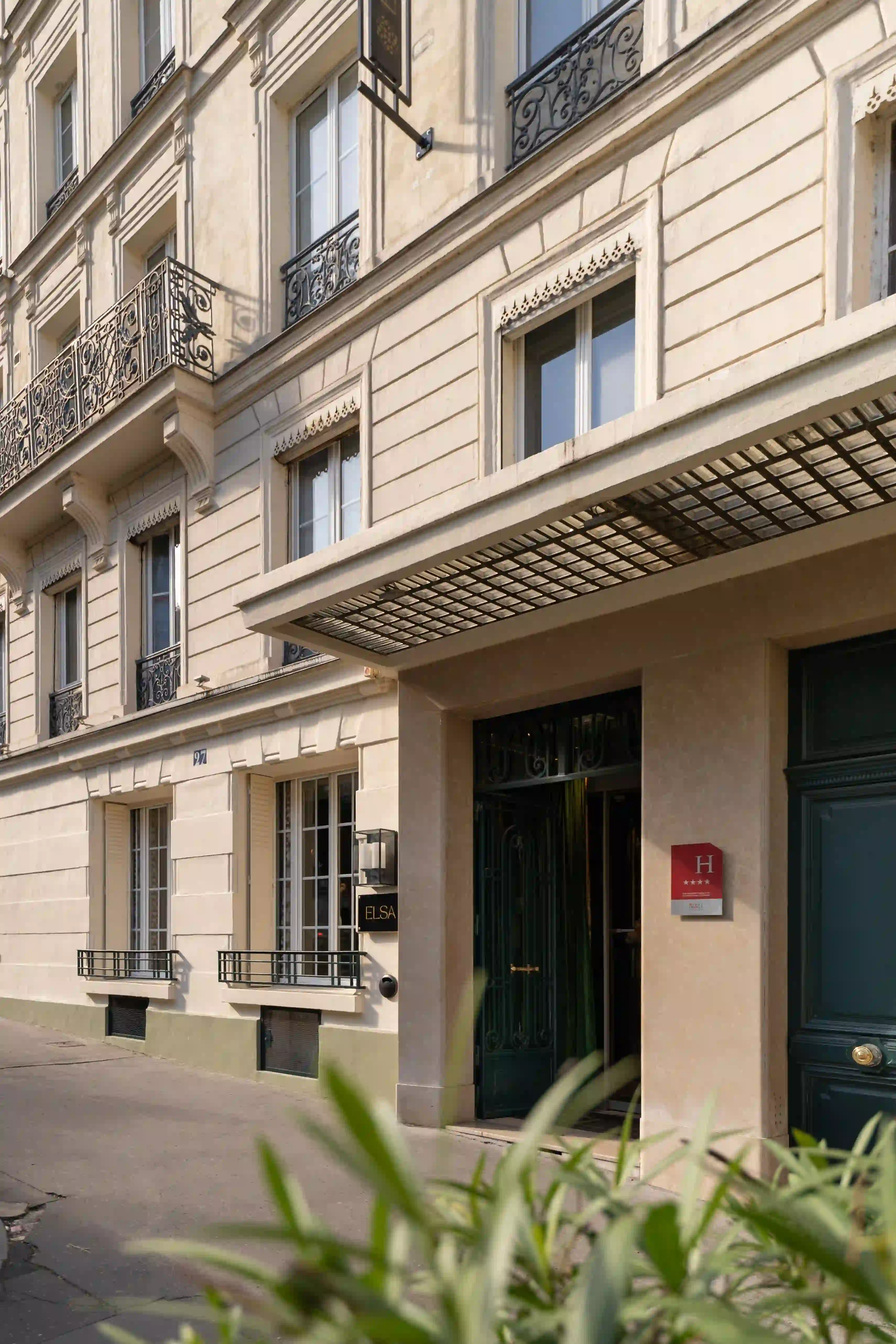 Fachada bege elegante de um edifício hoteleiro com varandas de ferro forjado e uma entrada com uma marquise metálica, uma placa vermelha indicando um hotel cinco estrelas e uma placa preta com o nome Elsa.