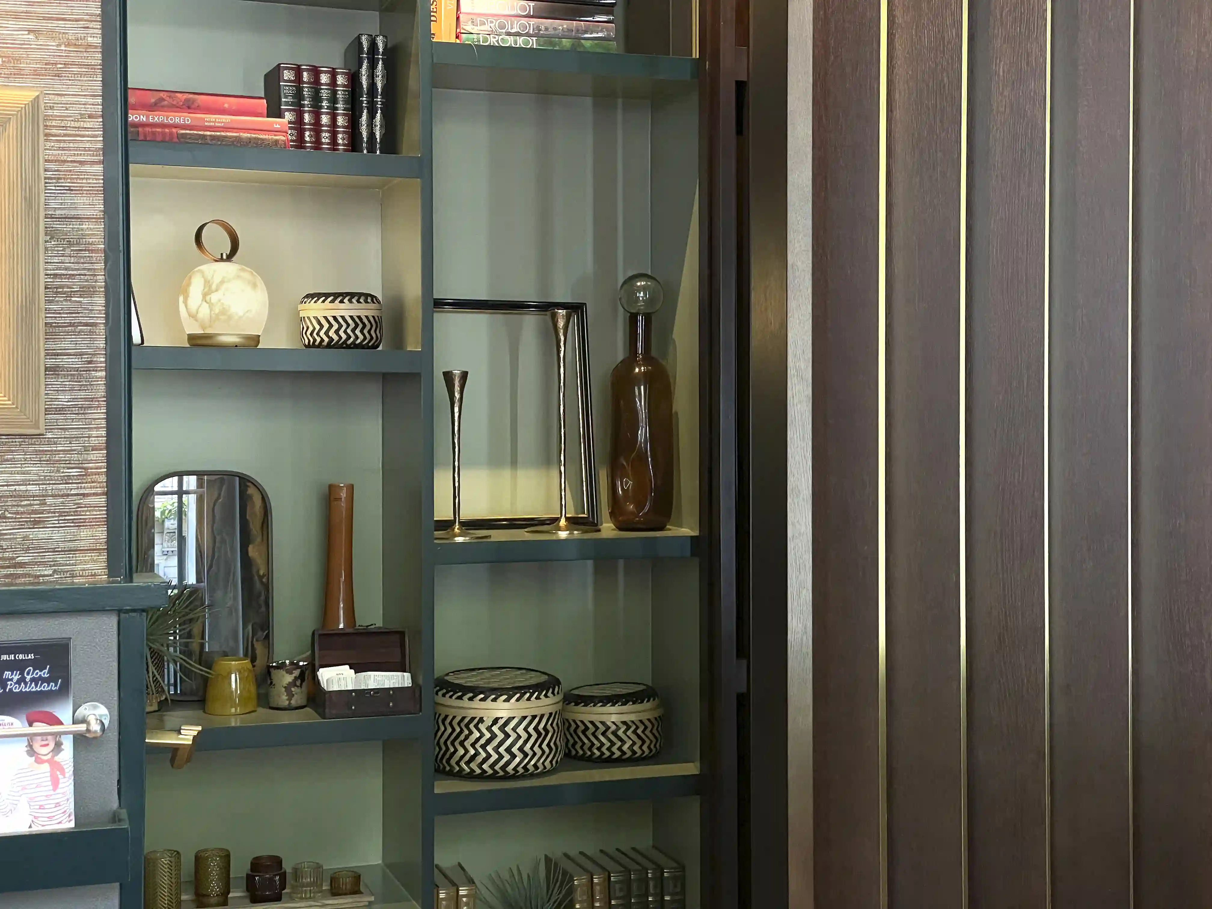 Prateleira decorativa com vários objetos, incluindo candeeiros, livros, cestos entrançados a preto e branco, vasos, uma moldura metálica e uma caixa de madeira, junto a uma parede com painéis de madeira castanha.