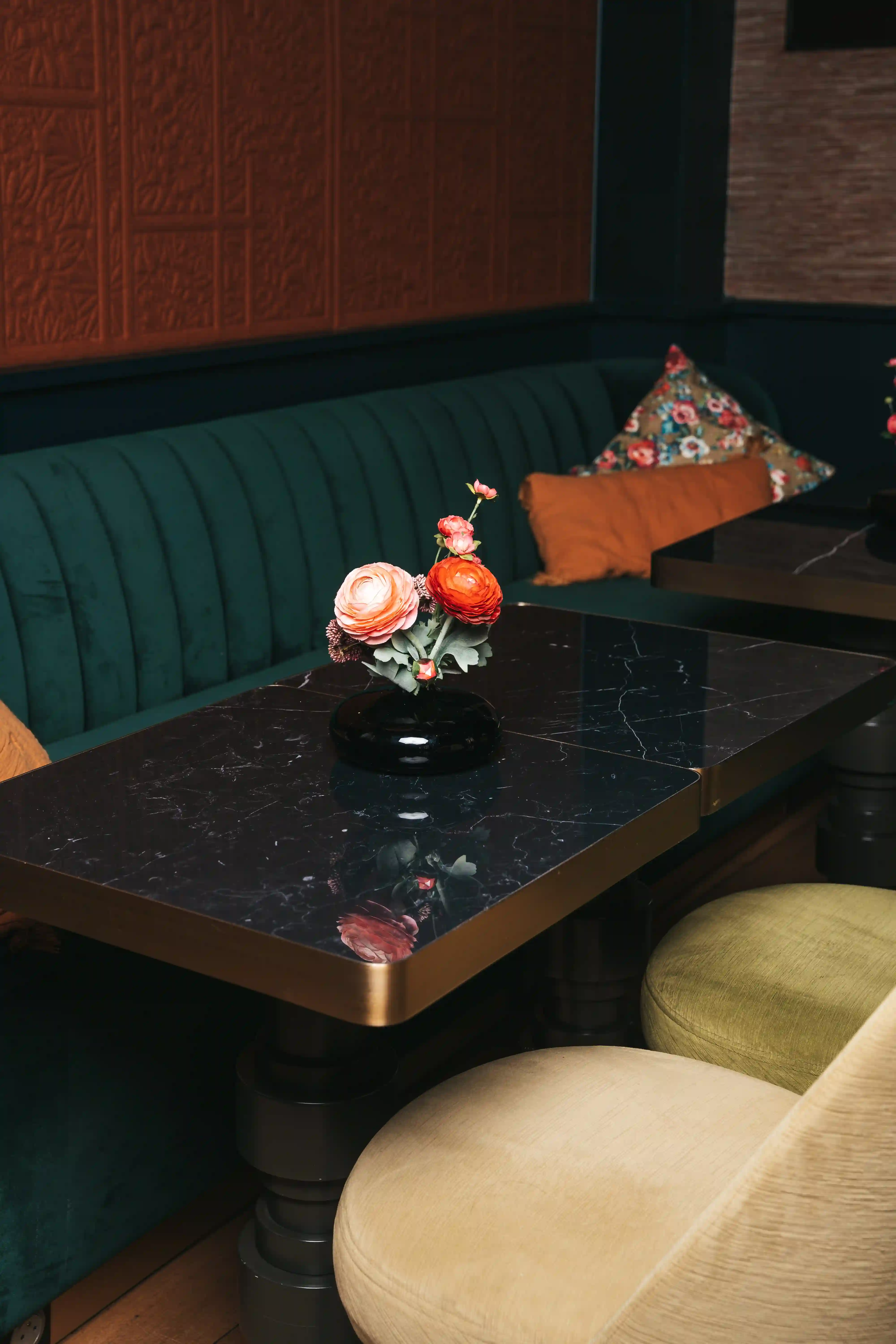 Mesa de mármore preta com um vaso preto contendo flores cor-de-rosa e laranja, rodeada por um banco verde e almofadas coloridas numa sala de estar elegante.