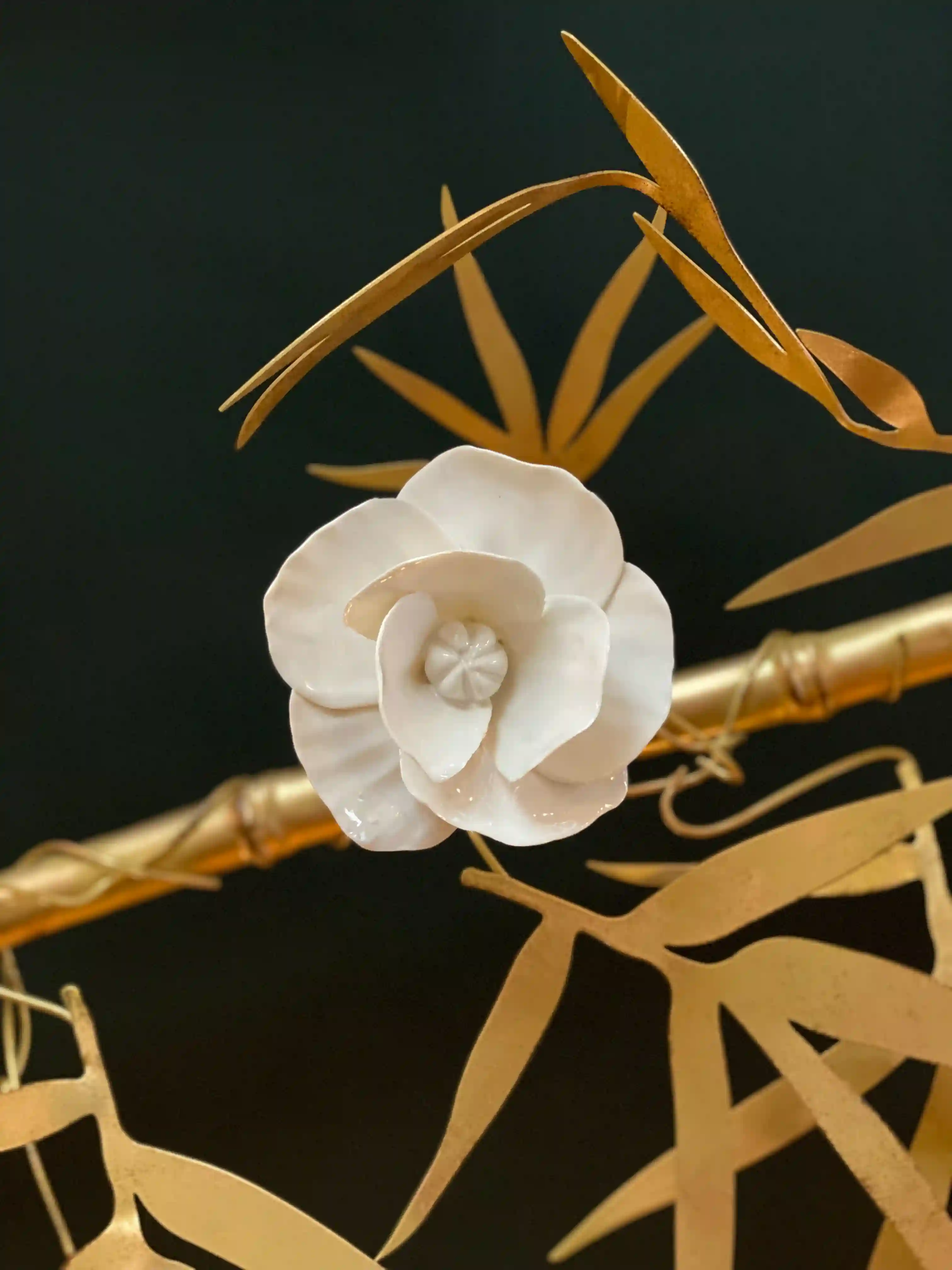 Rosa de porcelana branca delicada com folhas de metal dourado e haste dourada, sobre fundo preto.