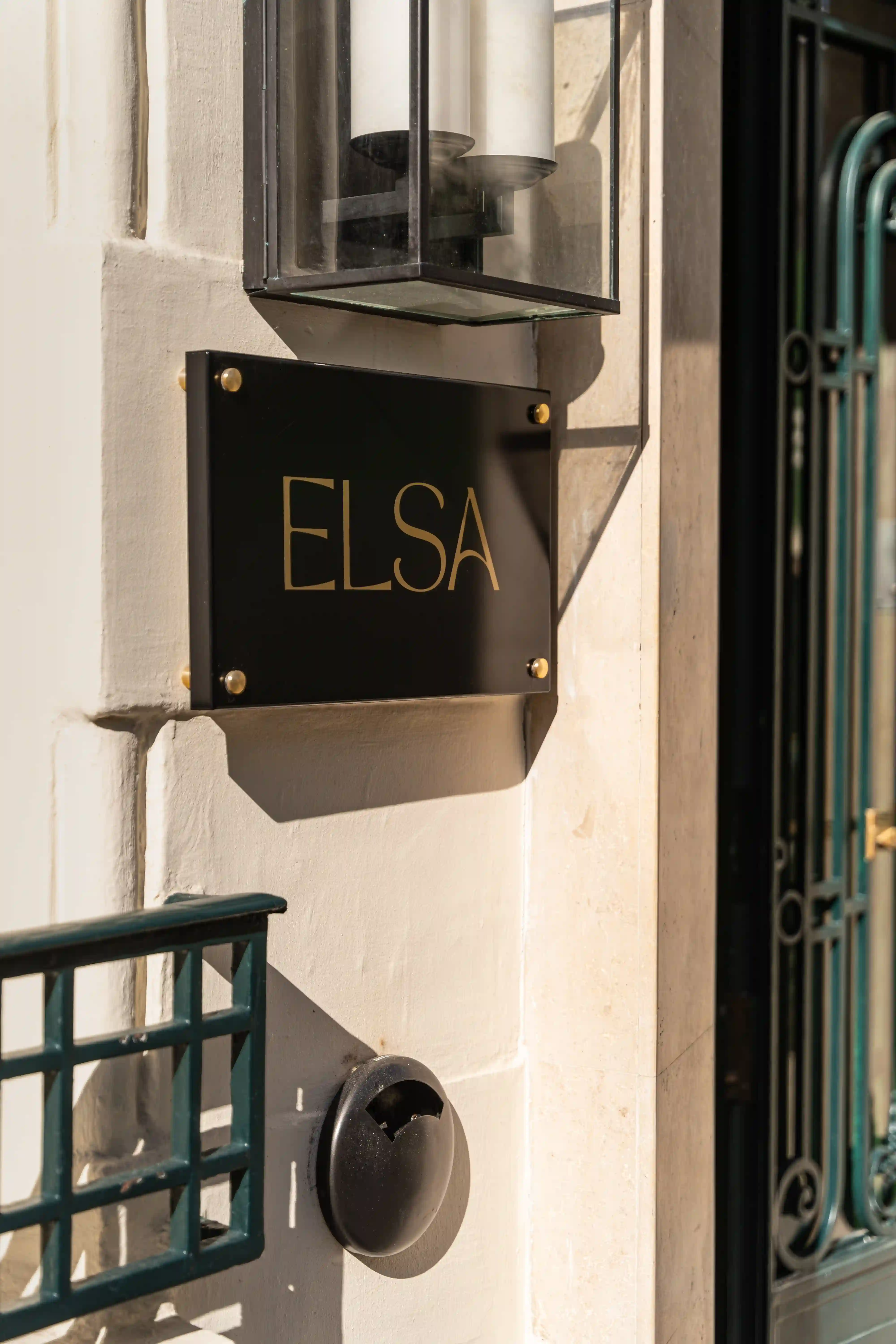 Plaque noire avec inscription dorée "ELSA" fixée sur un mur clair près d'une lampe murale en verre et d'une grille métallique verte, éclairée par le soleil.