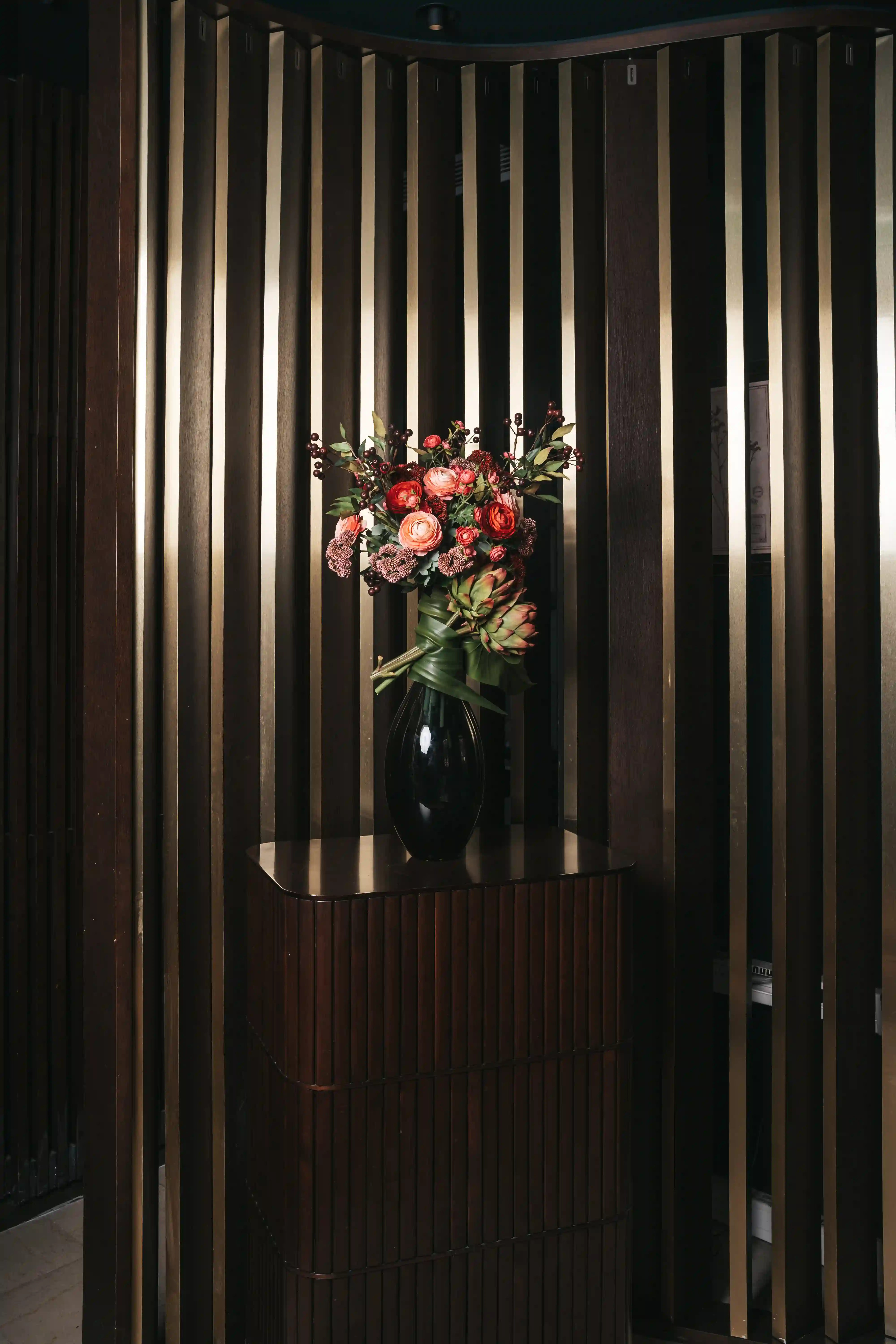 Bouquet de fleurs composé de roses, artichauts décoratifs, feuilles vertes, et baies rouges dans un vase noir placé sur une console en bois foncé devant un mur en lattes verticales sombres et dorées.