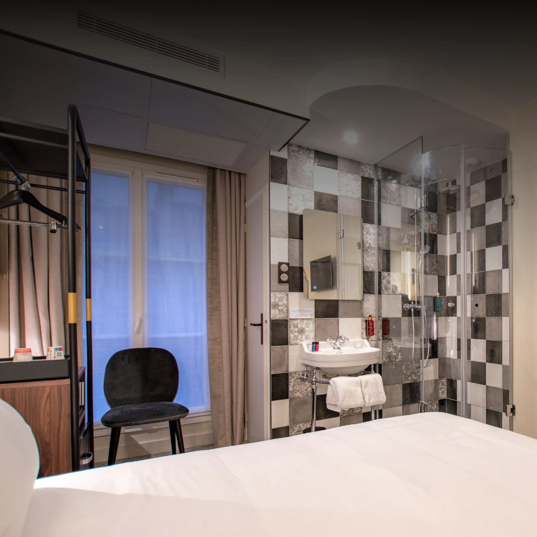 Quarto de hotel moderno com cama branca em primeiro plano, uma cadeira preta perto de uma janela com cortinas, um cabideiro de metal preto e um banheiro aberto com chuveiro de paredes de vidro e parede revestida com azulejos em xadrez preto, branco e cinza.