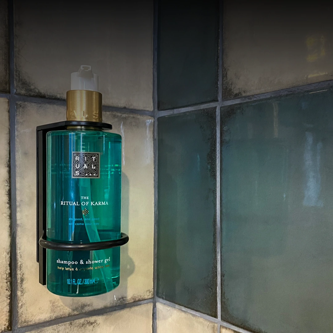 Dispensador de parede com gel de duche e champô The Ritual of Karma da marca Rituals, colocado sobre azulejos em tons de azul-esverdeado e bege.