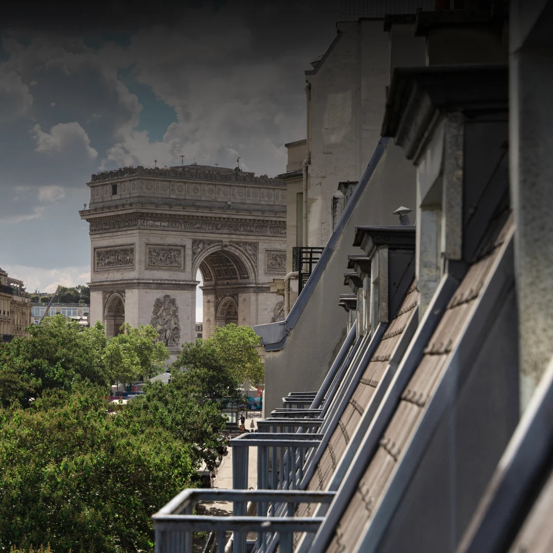 Vista do Arco do Triunfo em Paris, captada a partir de um terraço na lateral de um edifício, com telhados inclinados e árvores em primeiro plano sob um céu parcialmente nublado.