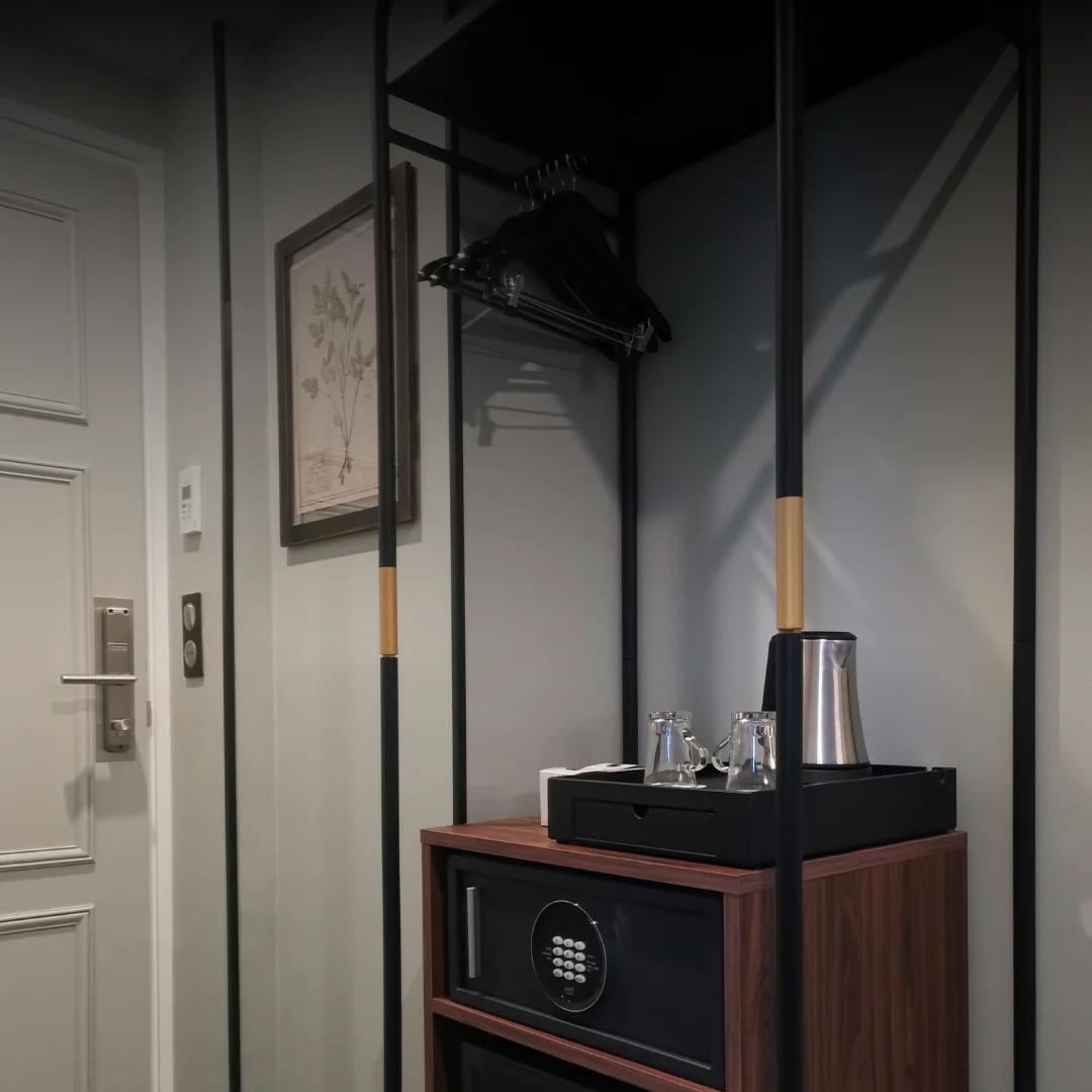 Canto de um quarto com um cabideiro preto com cabides, um cofre embutido num móvel de madeira com uma bandeja preta em cima contendo uma chaleira elétrica e duas chávenas de vidro, uma parede branca com um quadro decorativo e uma porta branca com fechadura eletrónica.