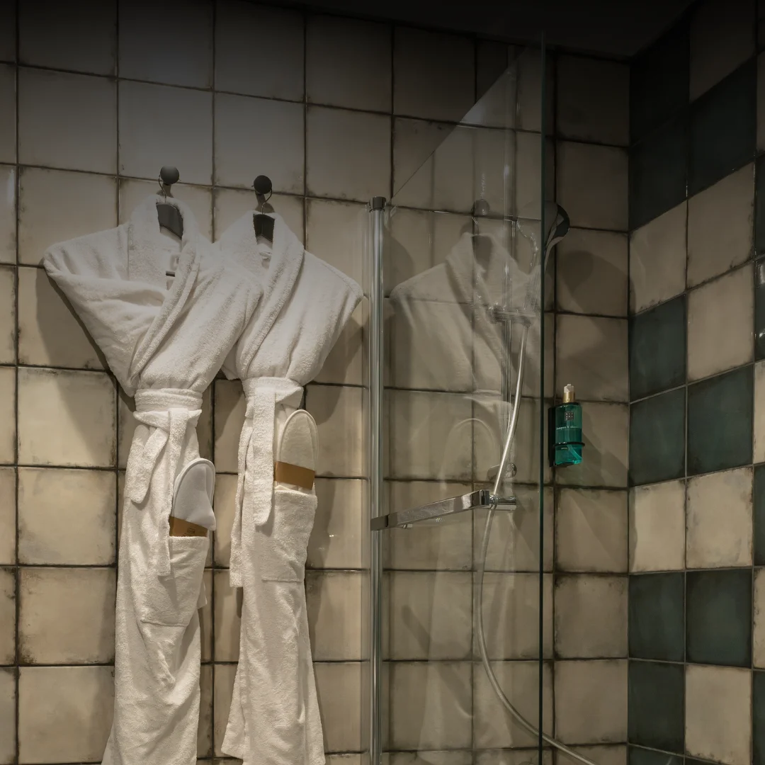 Dois roupões brancos pendurados em ganchos numa parede de azulejos ao lado de uma cabine de duche em vidro com um dispensador de sabonete verde fixado na parede.