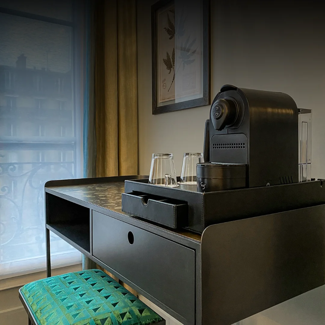 Máquina de café Nespresso preta sobre uma bandeja de couro com dois copos virados numa secretária preta e um banco com almofada verde sob a mesa, perto de uma janela com cortinas amarelo-esverdeadas e um quadro com desenho de folhas na parede.