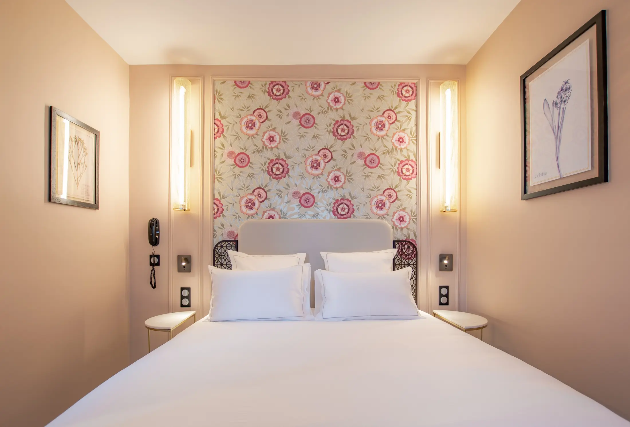 Elegant hotel room with a white double bed, four pillows, a grey headboard, and a pink and beige floral wall panel, illuminated by narrow wall lamps and decorated with two floral frames on beige walls.