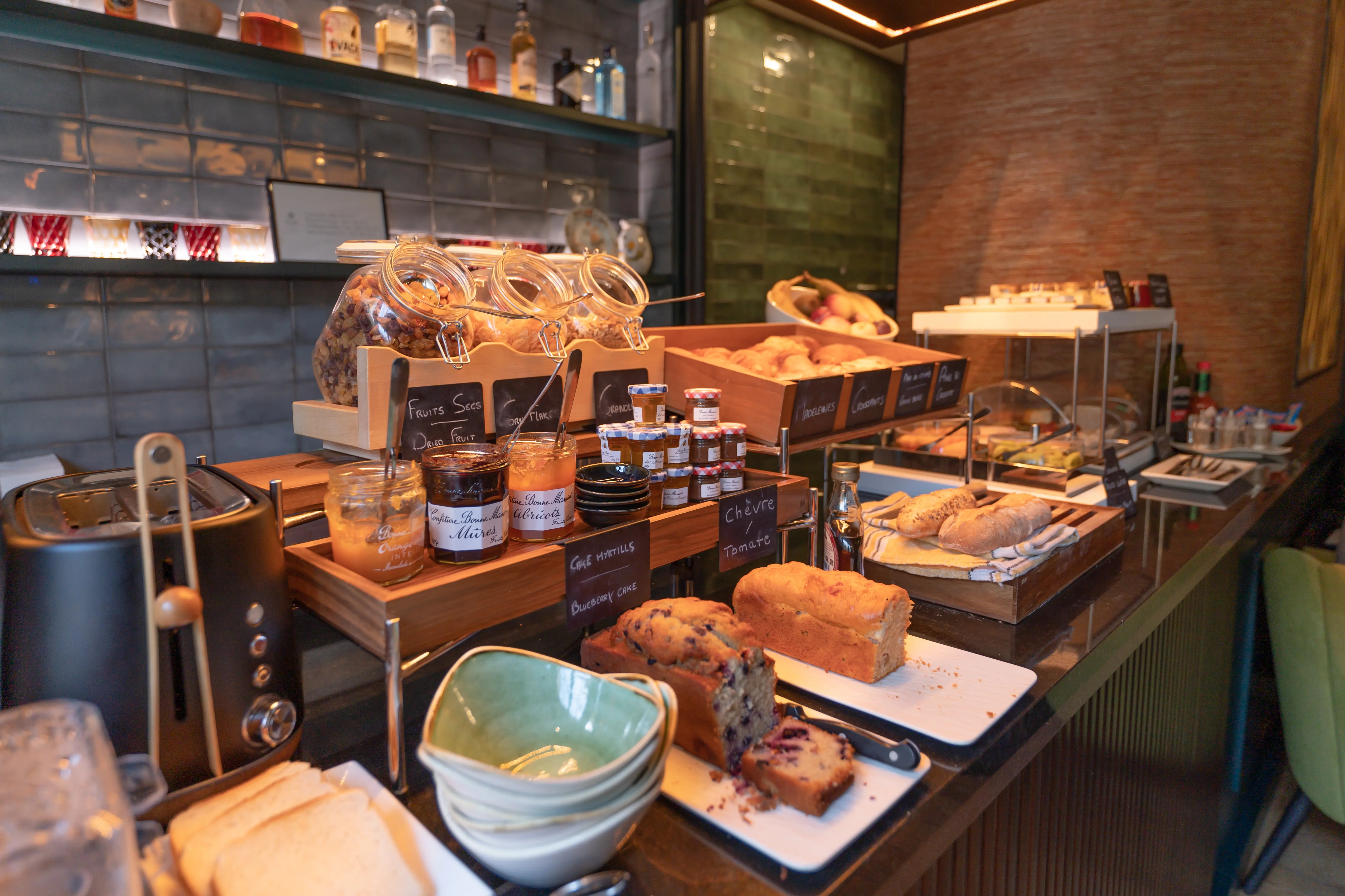 Breakfast buffet with bread, pastries, jams, dried fruits, cakes, stacked bowls, a black toaster, and bottles on a counter in a room with modern decor.