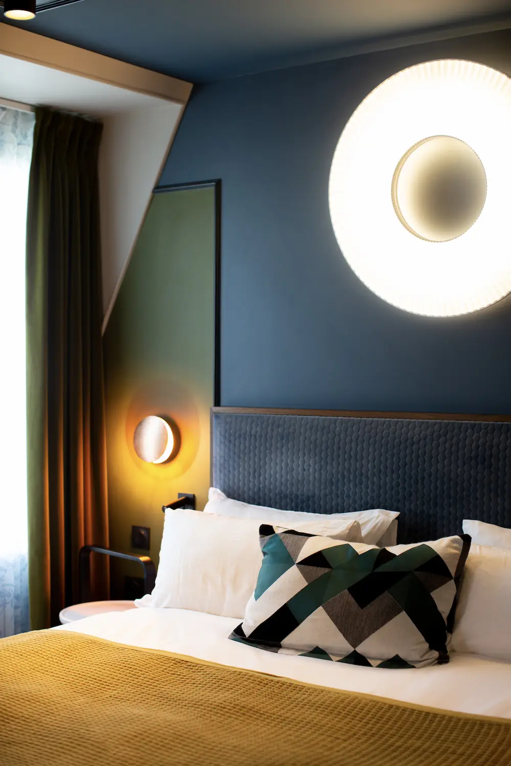Modern bedroom with patterned pillow, white pillows, yellow blanket, and circular wall lights.