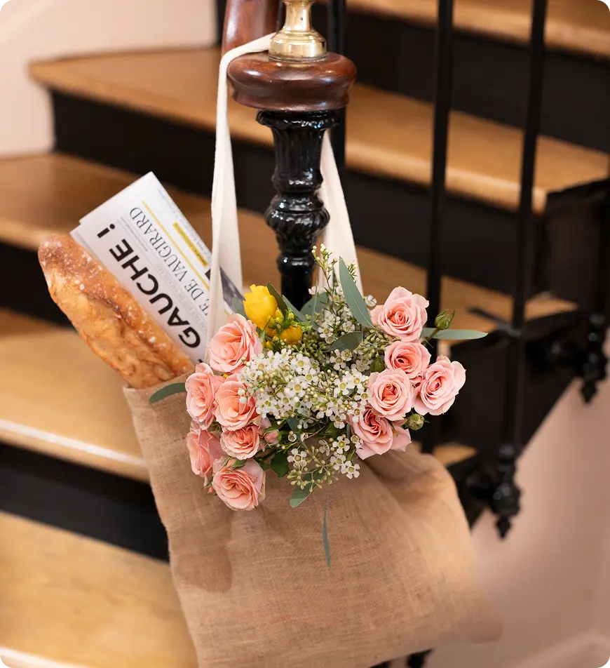 Interior com mesa preta de tampo brilhante e rebordo dourado, vaso preto com flores vermelhas e rosas, cadeira verde em primeiro plano, banco verde com almofadas florais e cor de laranja, e parede decorada com painéis cor de ferrugem.