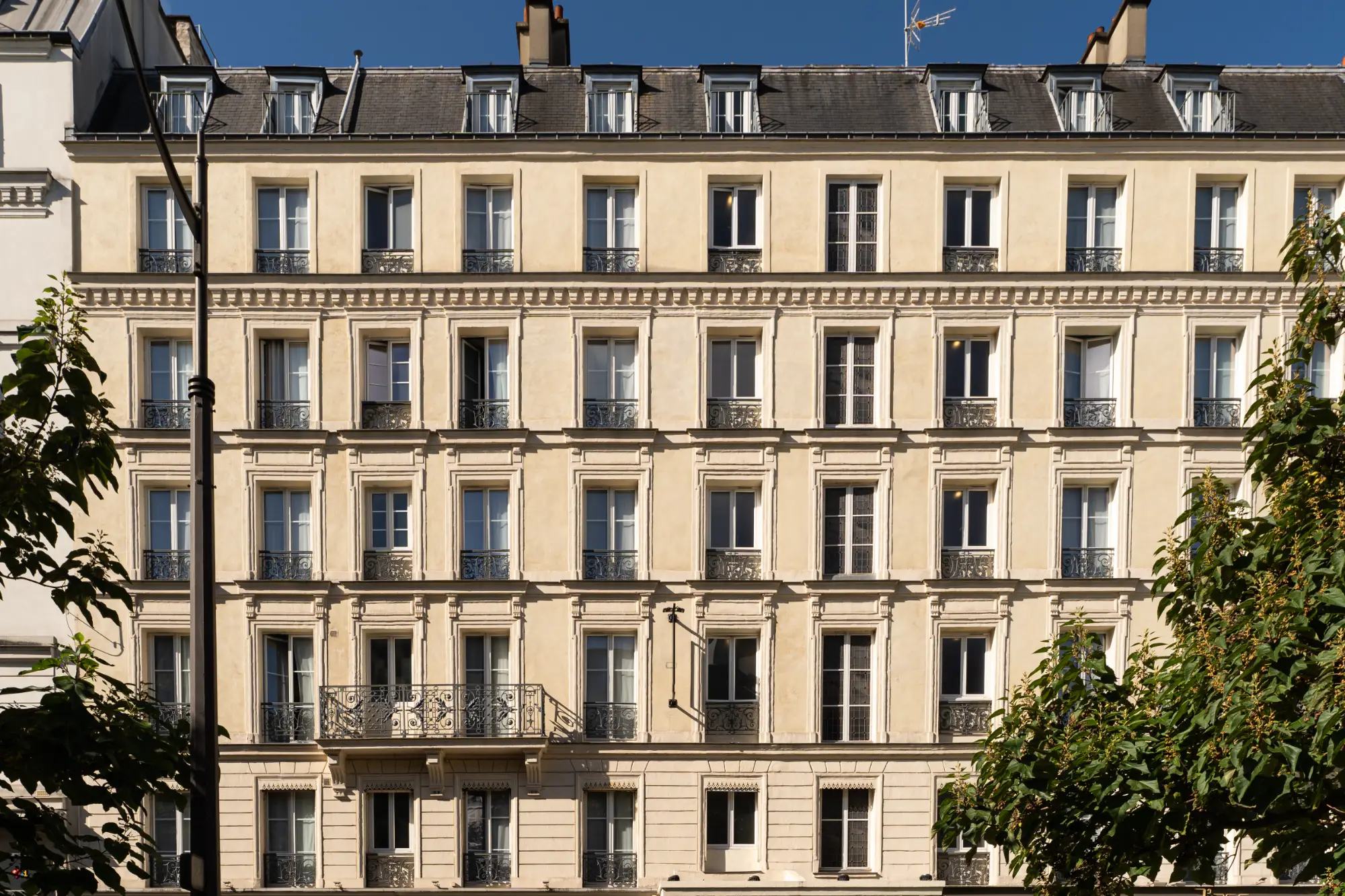 Fachada de um edifício parisiense clássico com vários andares de janelas com sacadas de ferro forjado, sob um céu azul claro, parcialmente emoldurada por árvores verdes