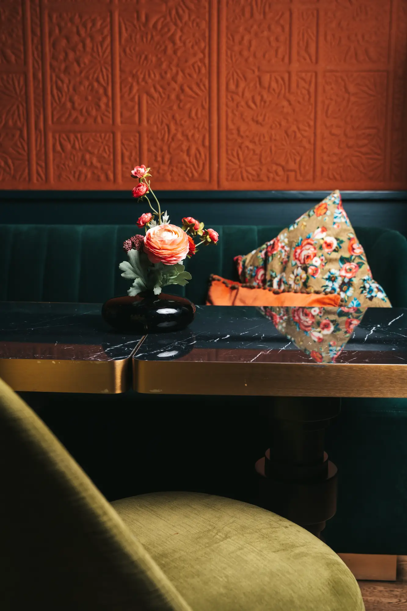 Intérieur avec table noire à plateau brillant et bord doré, vase noir avec fleurs rouges et roses, chaise verte au premier plan, banquette verte avec coussins fleuris et orange, mur orné de panneaux décoratifs rouille.