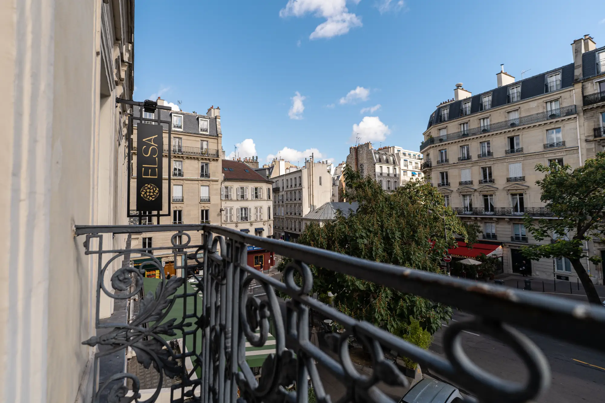 Vista a partir de uma sacada de ferro forjado de um hotel chamado Elsa sobre uma rua parisiense ladeada por árvores e prédios haussmannianos sob um céu azul com algumas nuvens