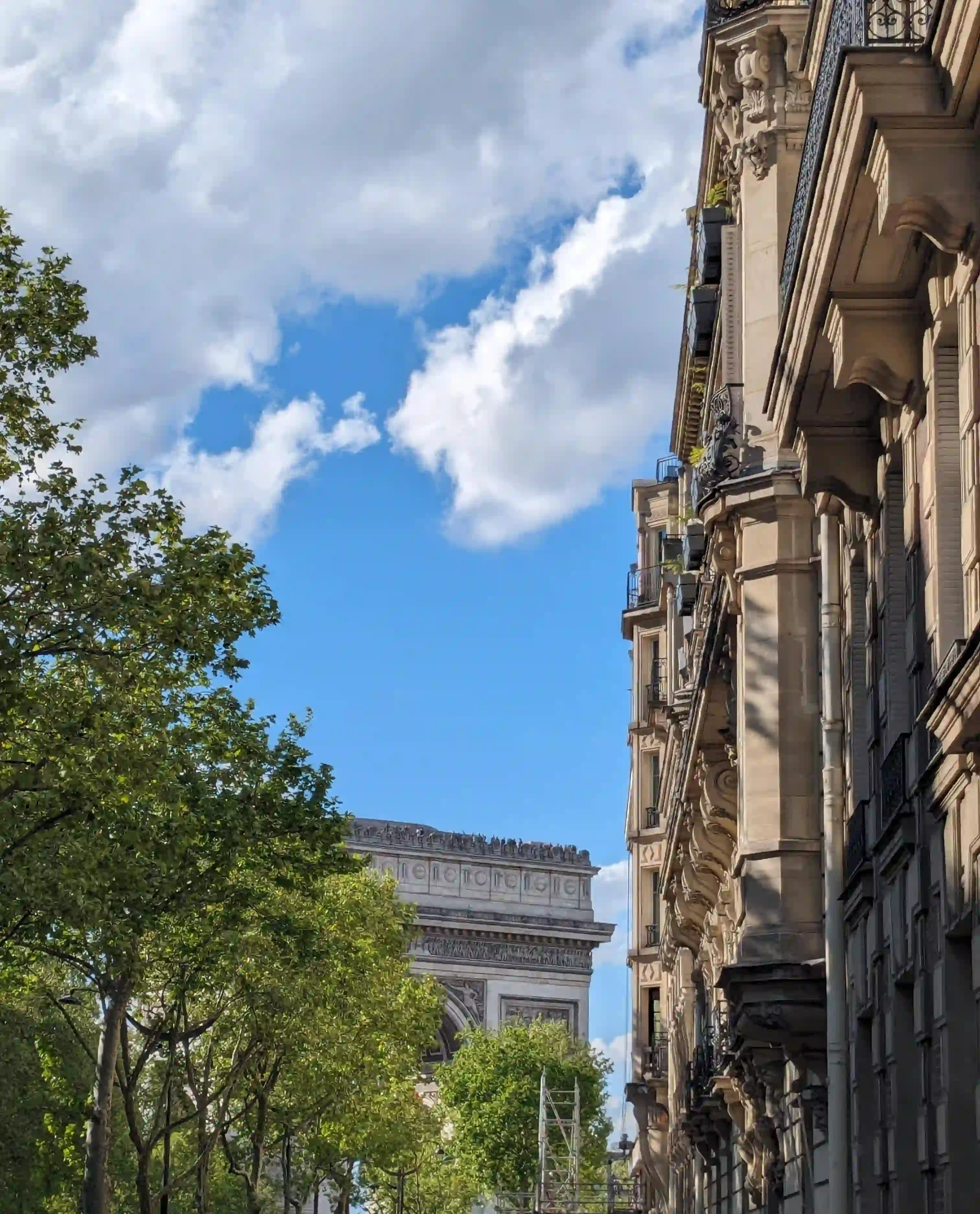 Partial view of the Arc de Triomphe in Paris between green trees on the left and a Haussmann-style building on the right under a blue sky with a few white clouds