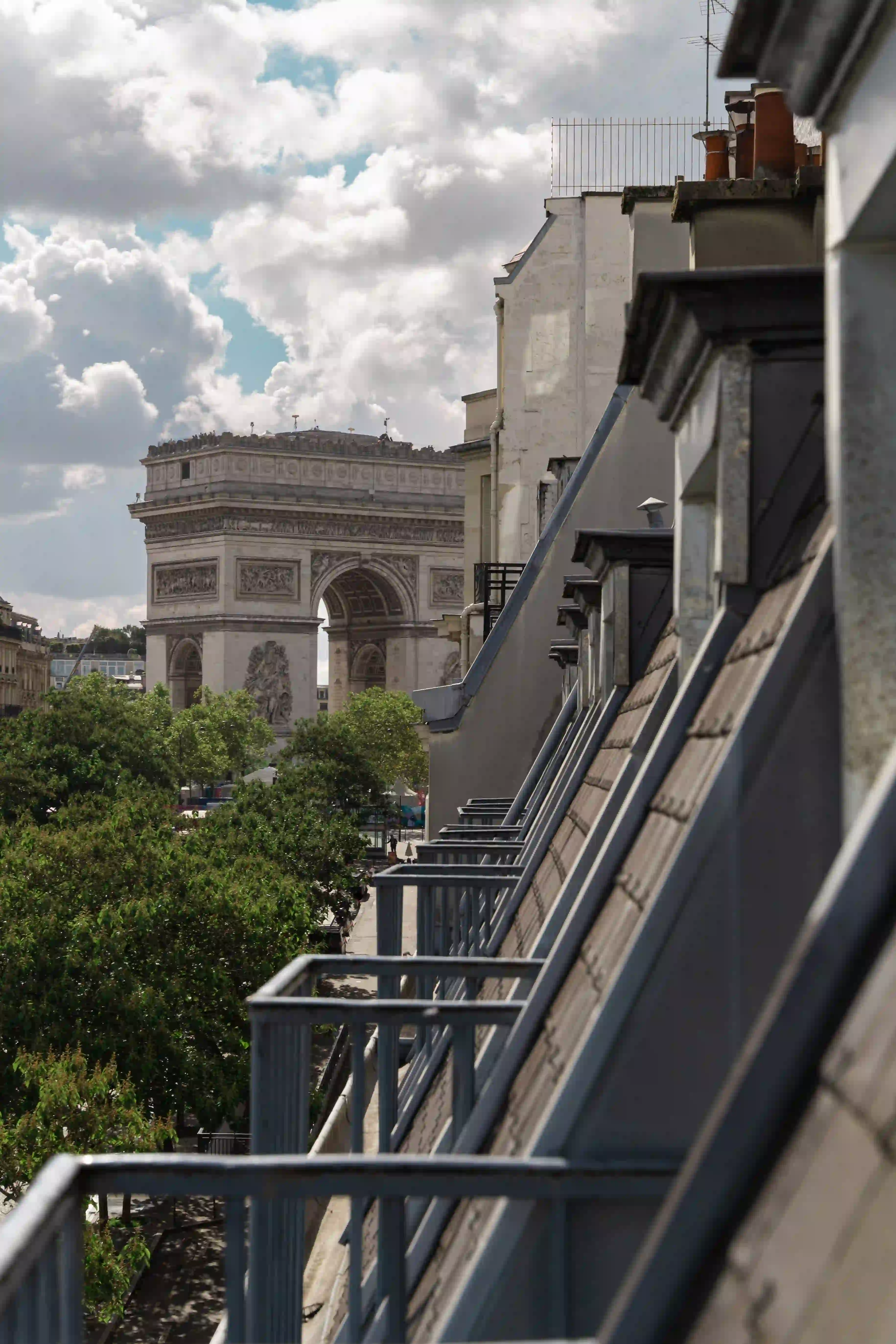 Vue depuis un toit parisien montrant plusieurs fenêtres mansardées en enfilade à droite, avec l'Arc de Triomphe visible au loin à gauche, sous un ciel partiellement nuageux.