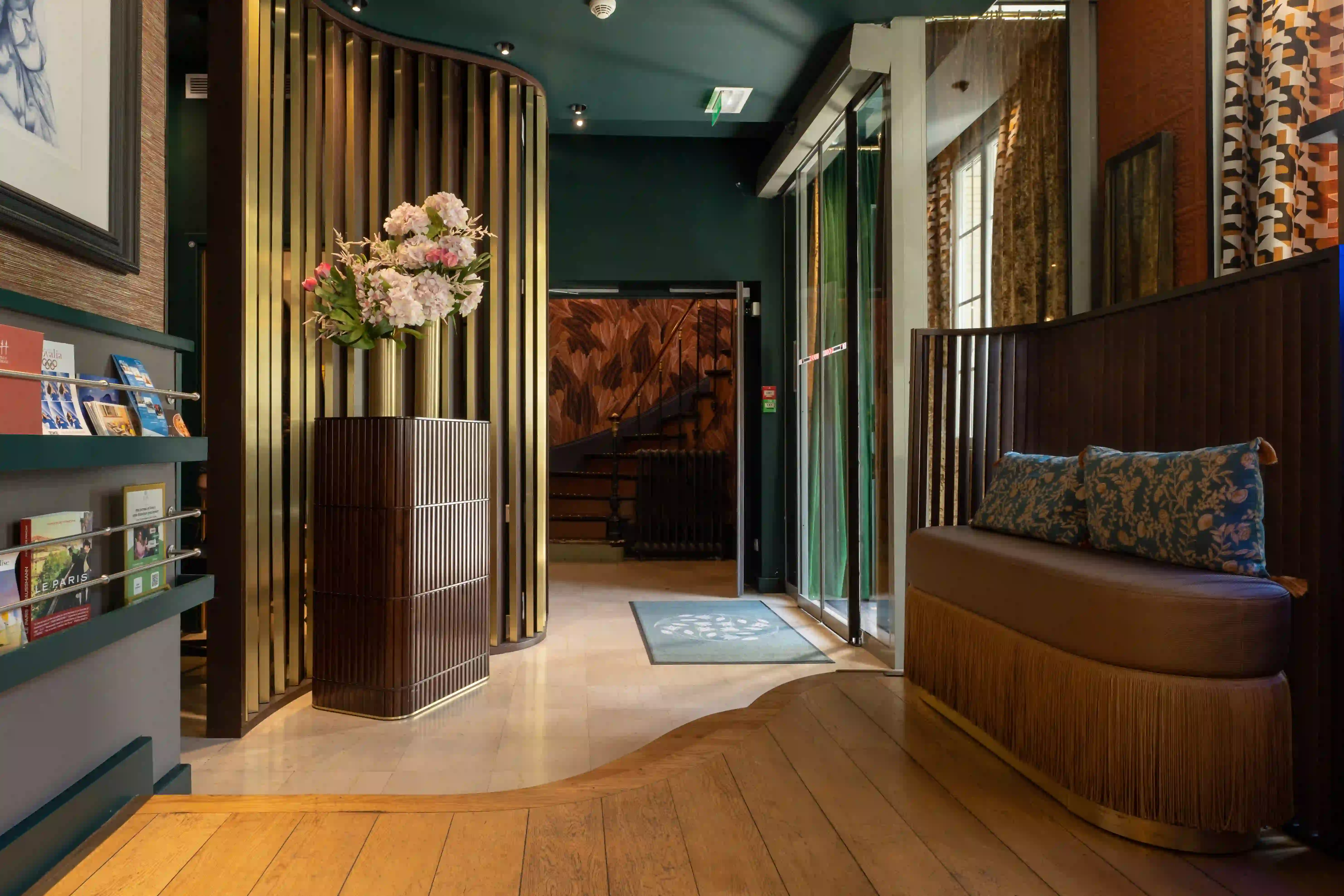 Hall d'entrée d'hôtel élégant avec sol en bois ondulé, canapé beige orné de coussins à motifs floraux bleus, grandes fenêtres avec rideaux, une cloison en bois et une table centrale arrondie portant un vase doré avec des fleurs blanches et roses, murs foncés et escalier visible à l'arrière.