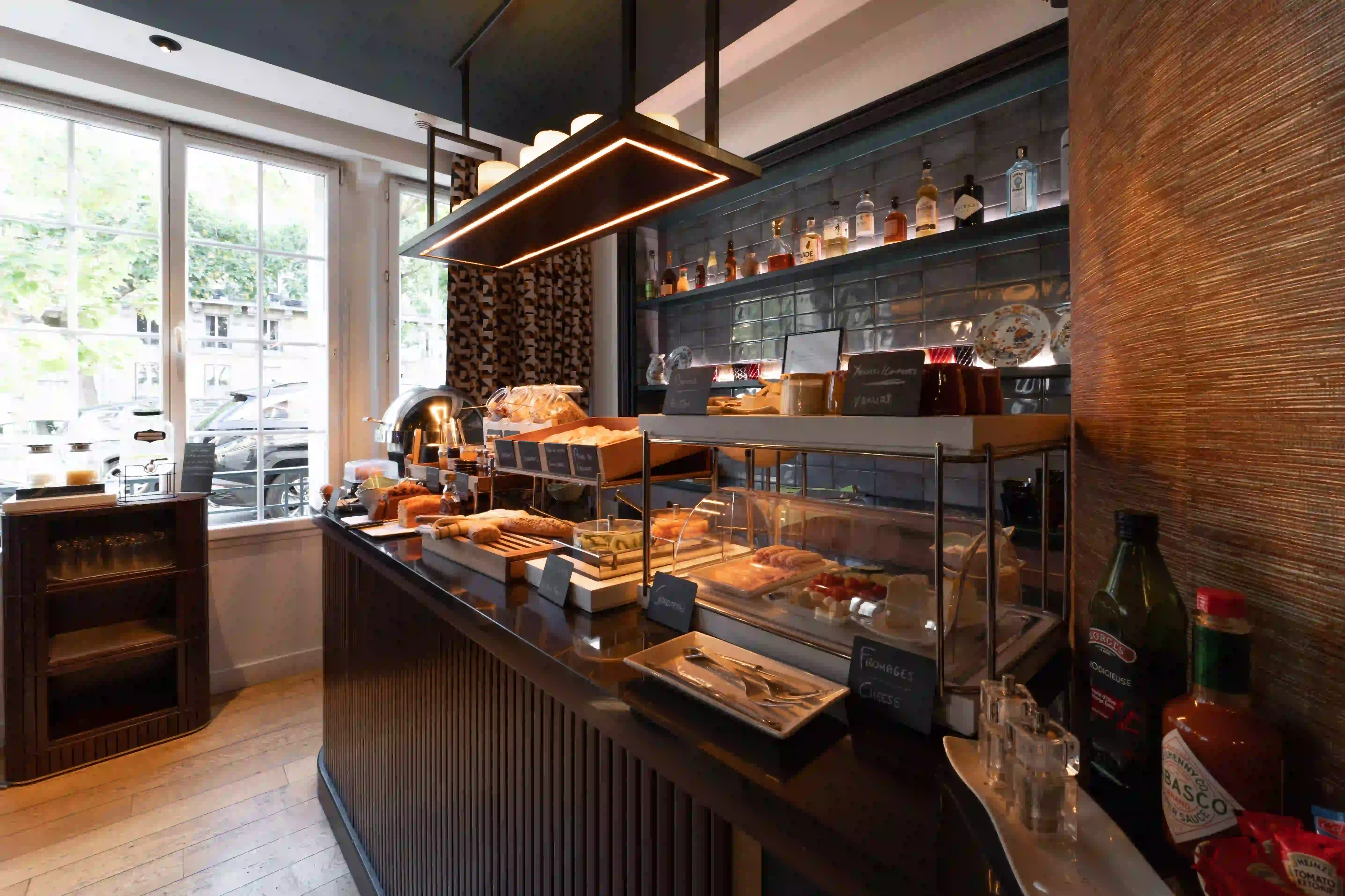 Breakfast buffet counter in a warm setting with pastries, breads, cheeses, yogurts, condiments, and bottles of spirits on illuminated shelves in the background.