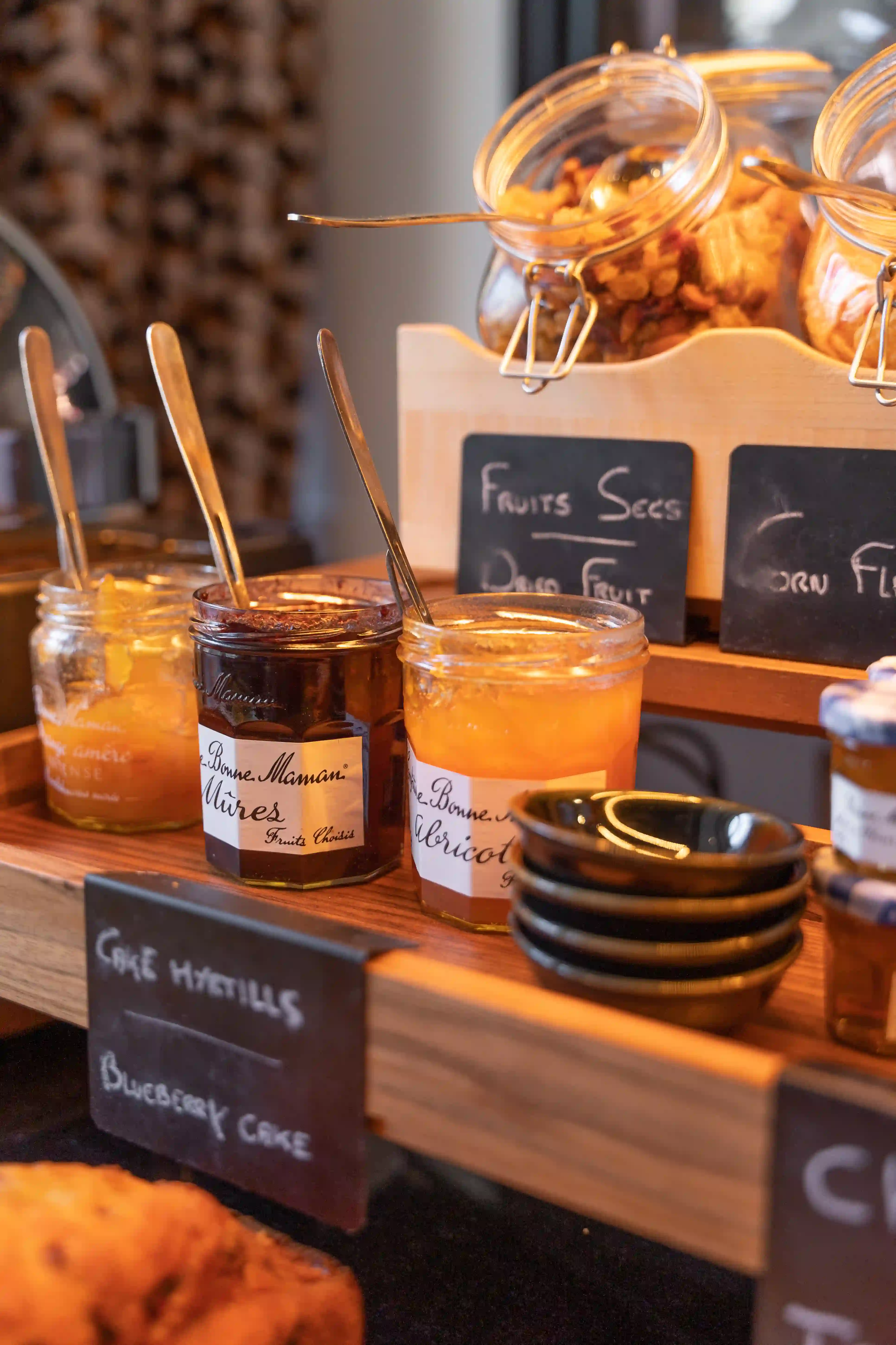 Three Bonne Maman jam jars with spoons, a jar of blackberries, a jar of apricots and another blurred jar, placed on a wooden shelf with chalk-written labels indicating dried fruits and blueberry cake