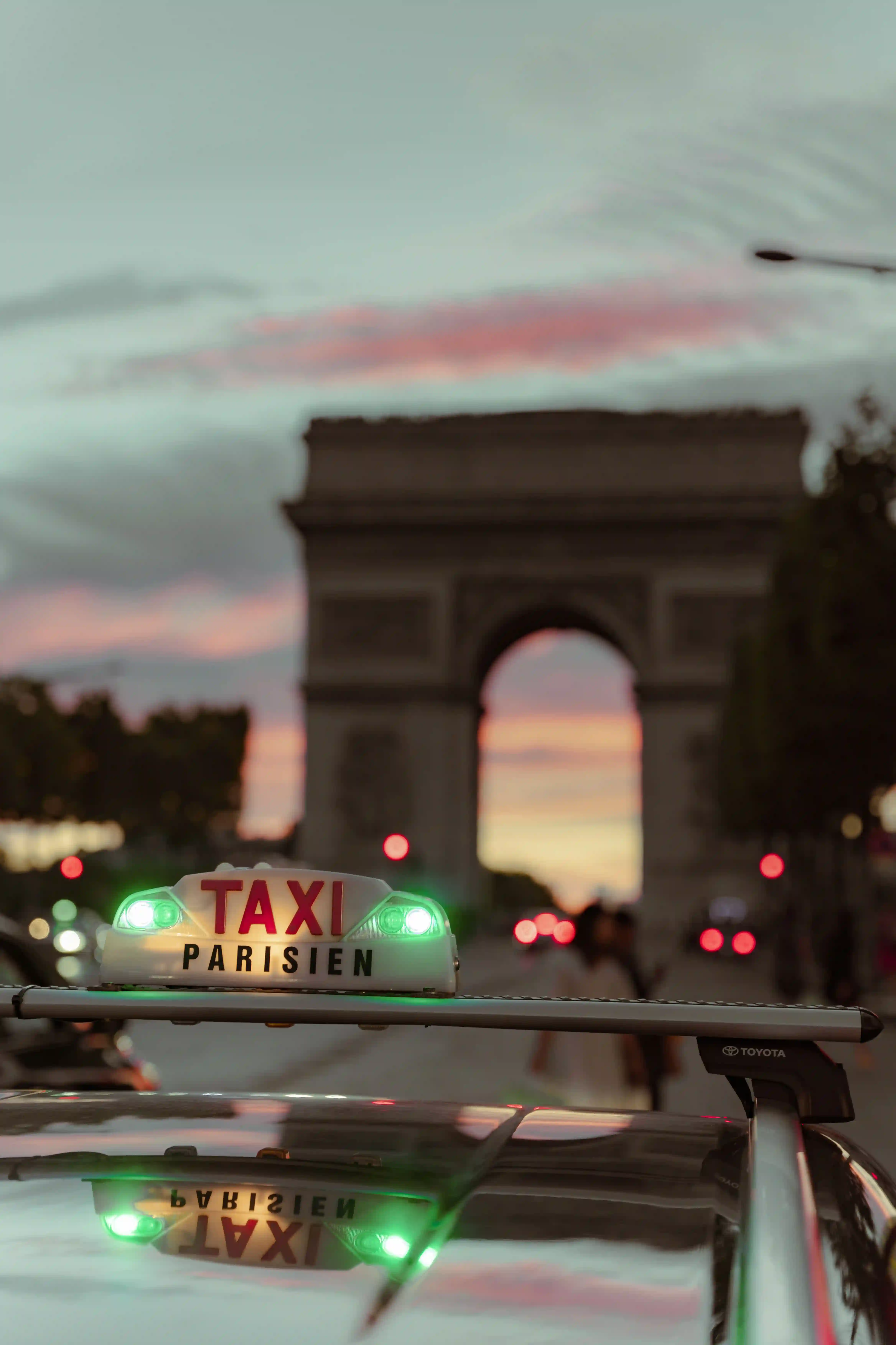 A Parisian taxi light sign lit in green mounted on the roof of a Toyota car, with the blurred Arc de Triomphe in the background at sunset