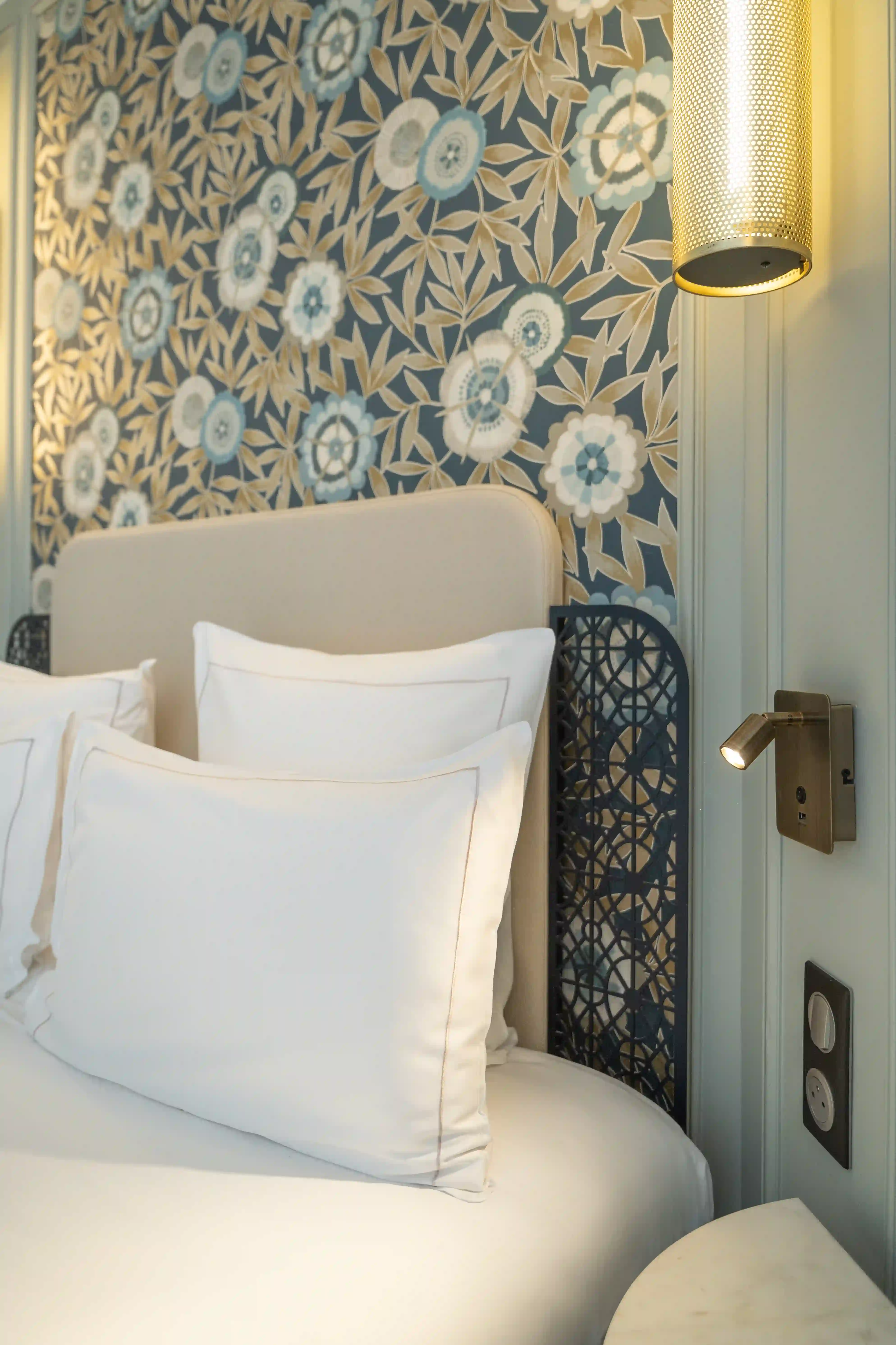 Détail de tête de lit beige avec deux grands oreillers blancs et un coussin rectangulaire blanc, devant un mur à motif floral bleu, gris et beige, éclairé par une lampe murale cylindrique dorée et une petite lampe de lecture orientable.