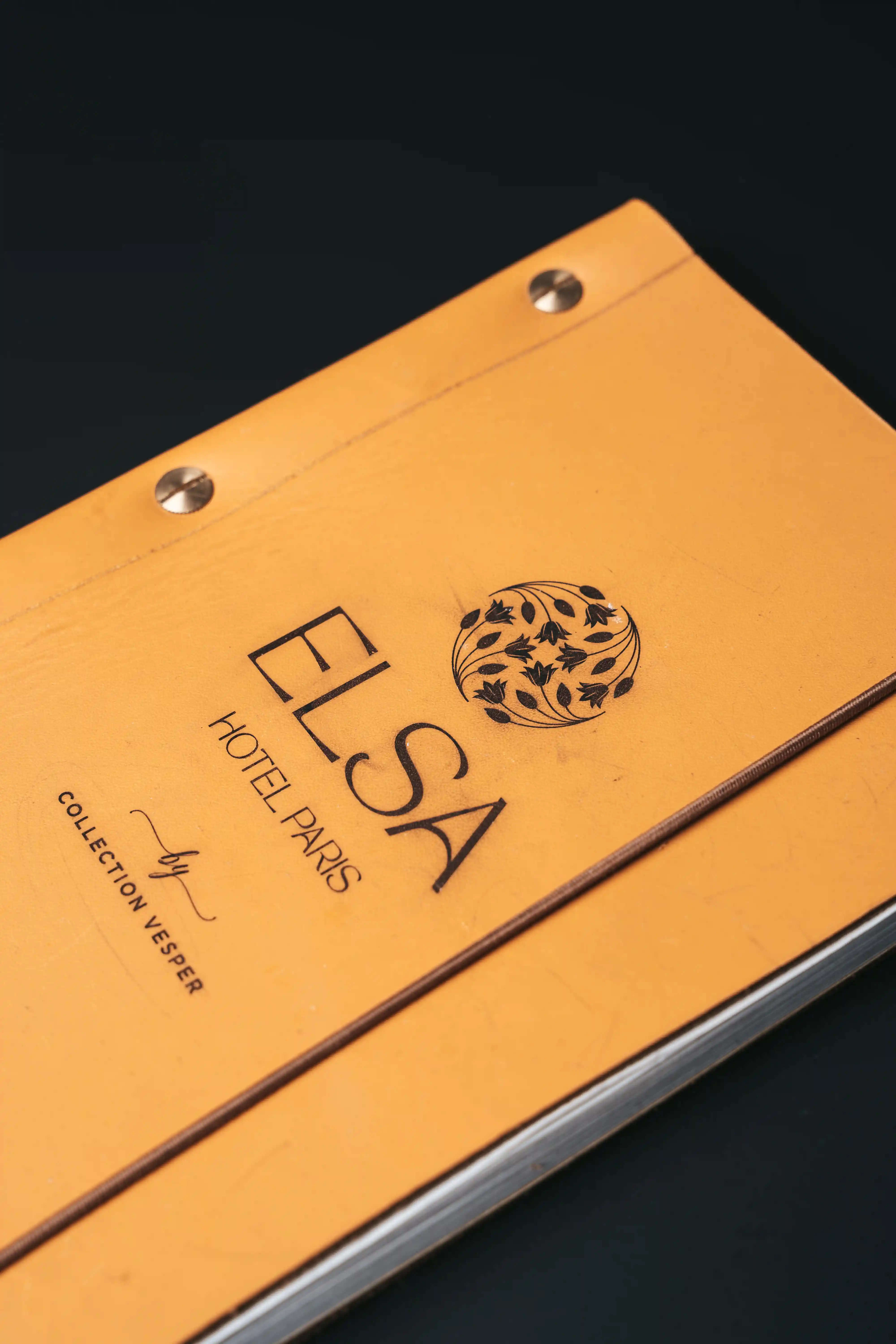 Yellow leather folder with the Elsa Paris Collection Vesper hotel logo and text, resting on a black surface.