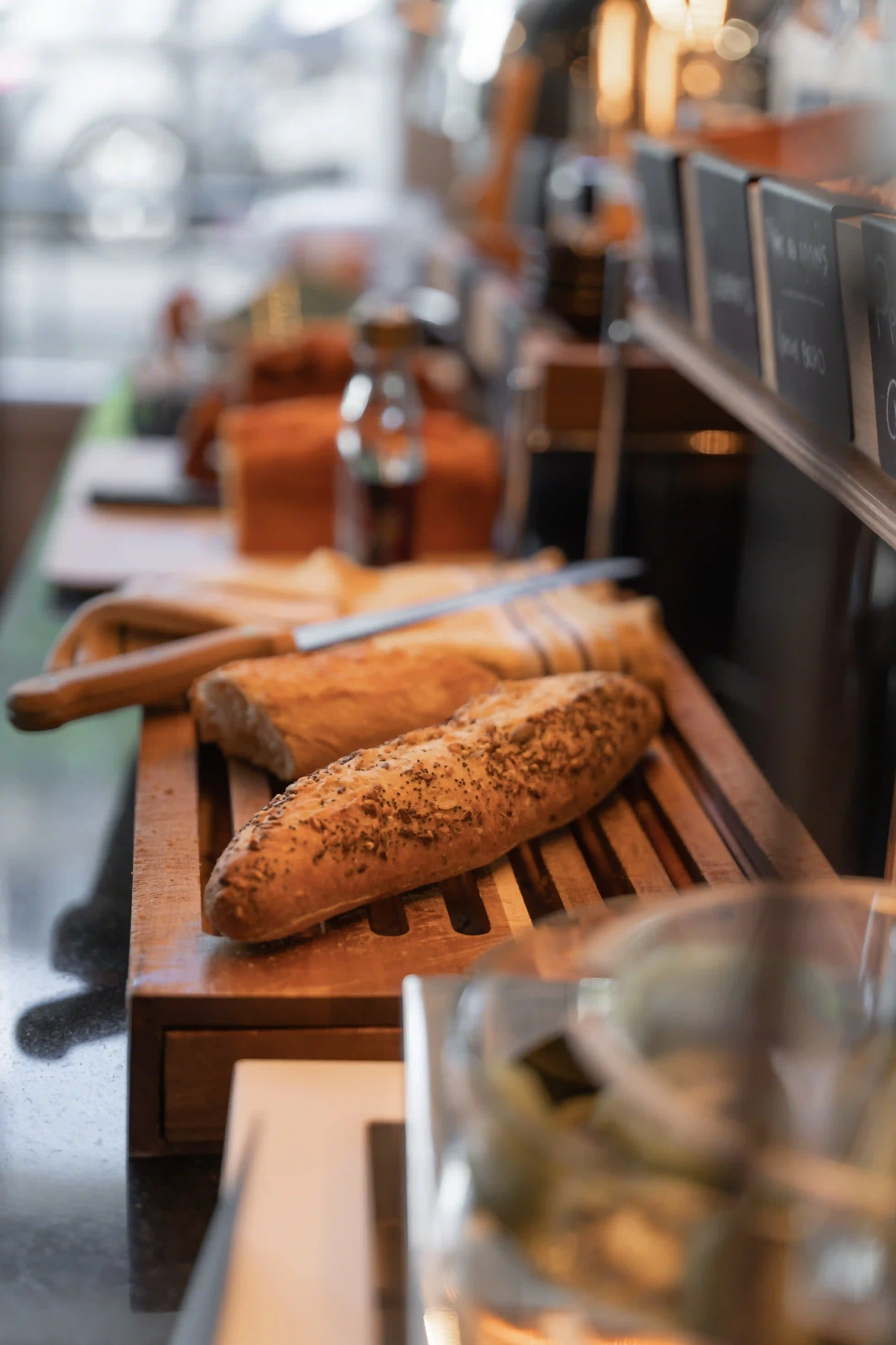 Frische Baguettes auf einem Holzbrett mit einem Messer, vor einer unscharfen Bäckerei- oder Küchenkulisse im Hintergrund