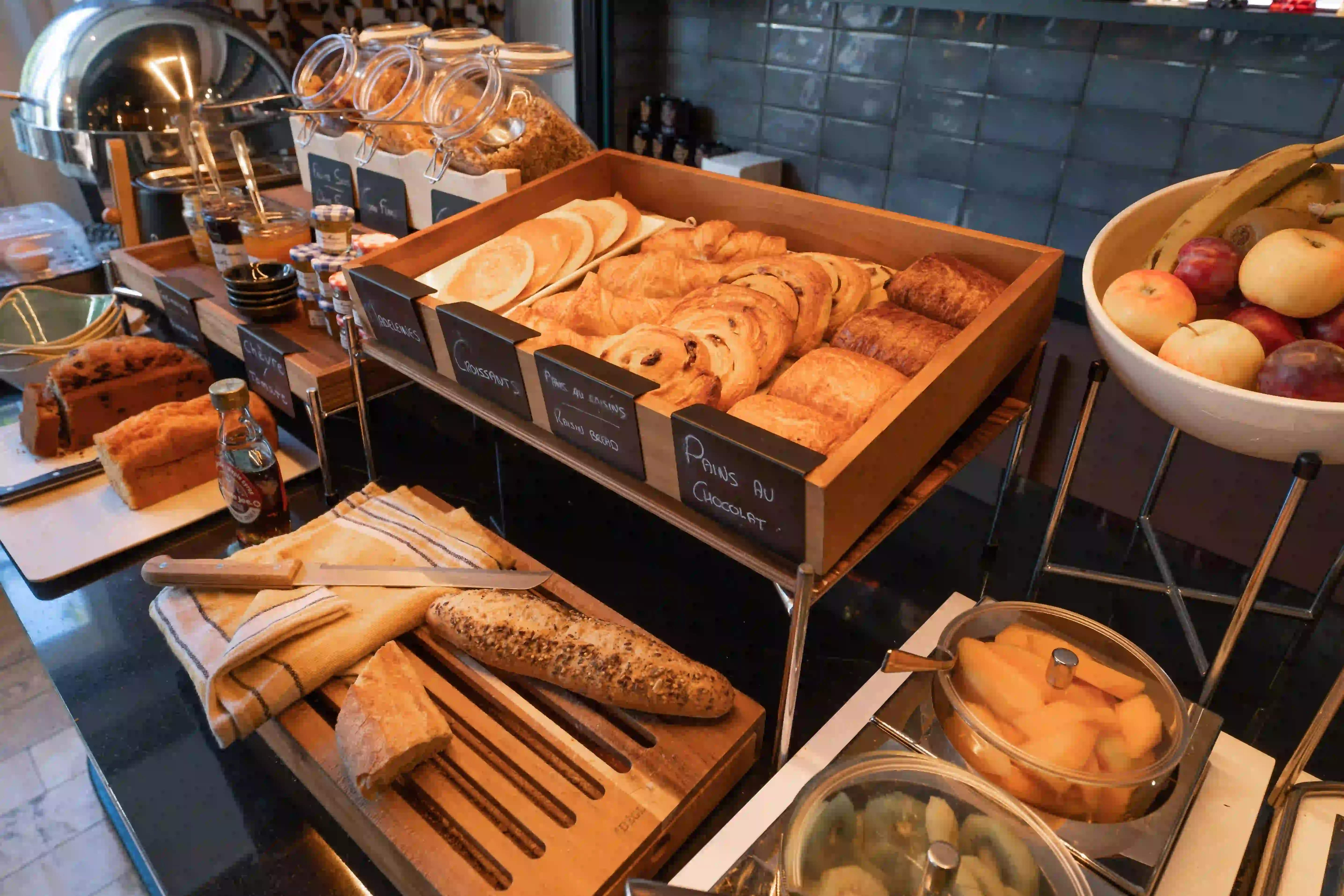 Frühstücksbuffet mit einer Auswahl an Feingebäck, darunter Crêpes, Croissants, Rosinenschnecken, Schokocroissants, geschnittene Baguettes, Marmeladen, Müsli, frische Früchte in einer Schale und geschnittene Früchte in Schüsseln mit Deckeln