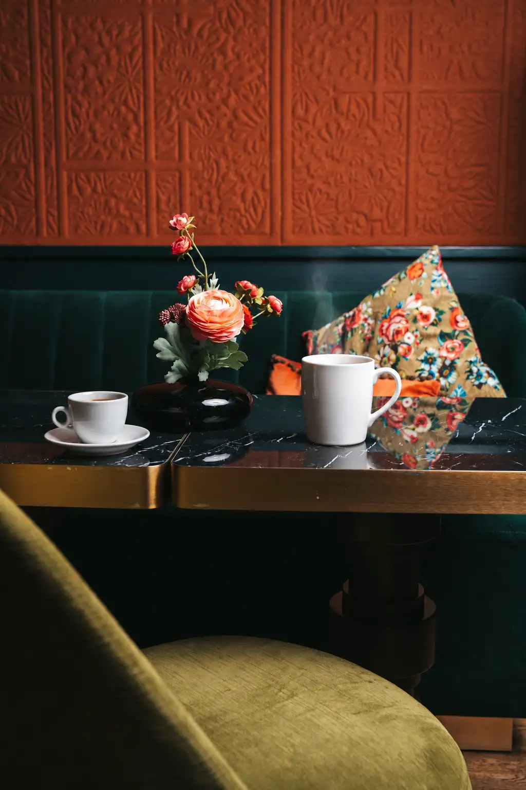 Ein schwarzer Marmortisch mit goldenem Rand, darauf eine Espressotasse auf einer Untertasse, eine dampfende weiße Tasse und eine schwarze Vase mit rosa und roten Blumen; im Hintergrund ein dunkelgrünes Sofa und ein buntes Blumenkissen vor einer strukturierten roten Wand.