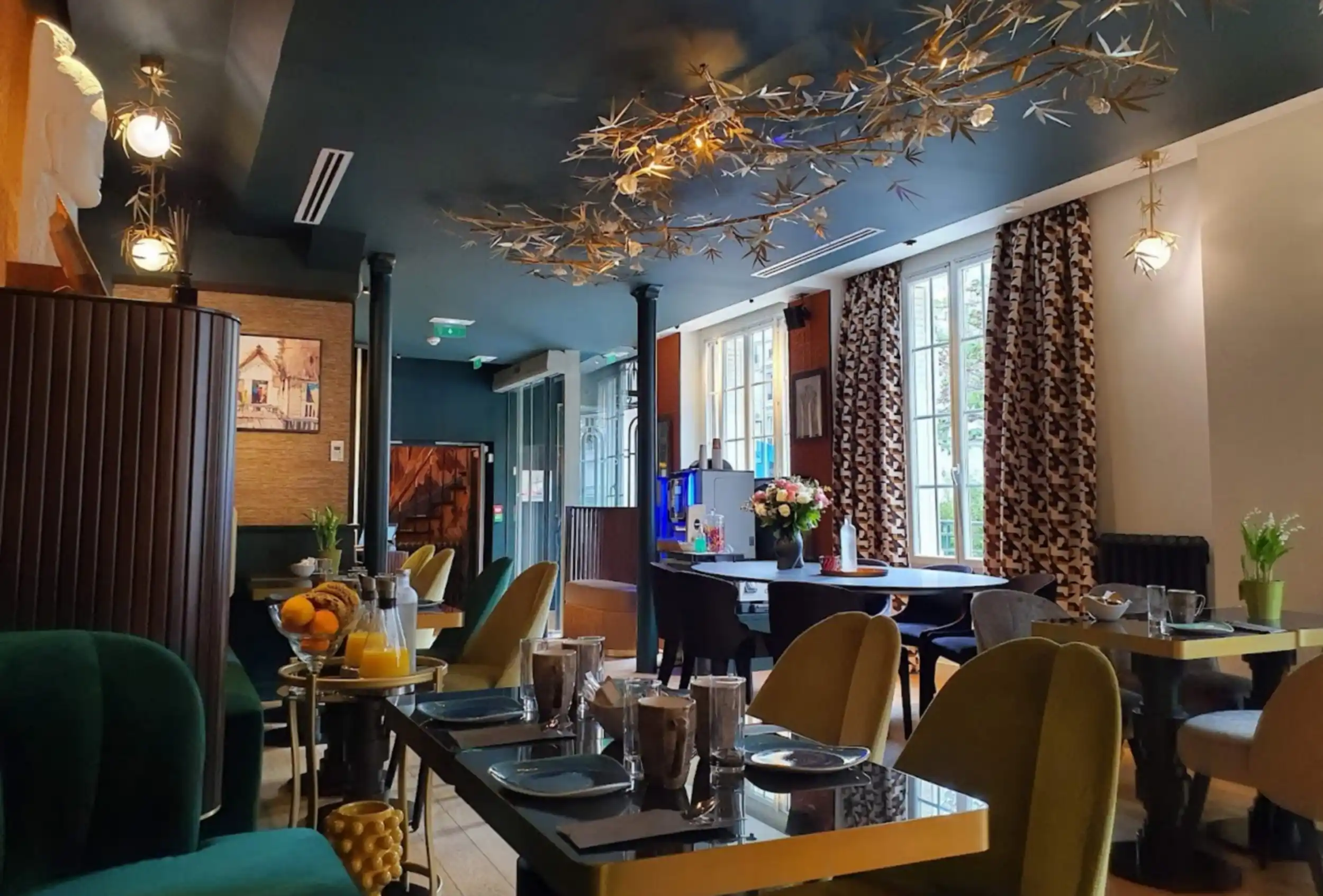 Elegantes Restaurant mit gedeckten Tischen mit Geschirr und Gläsern, grünen und gelben Stühlen, großen Fenstern mit gemusterten Vorhängen und einer dekorativen Hängeleuchte in Form goldener Zweige an der Decke