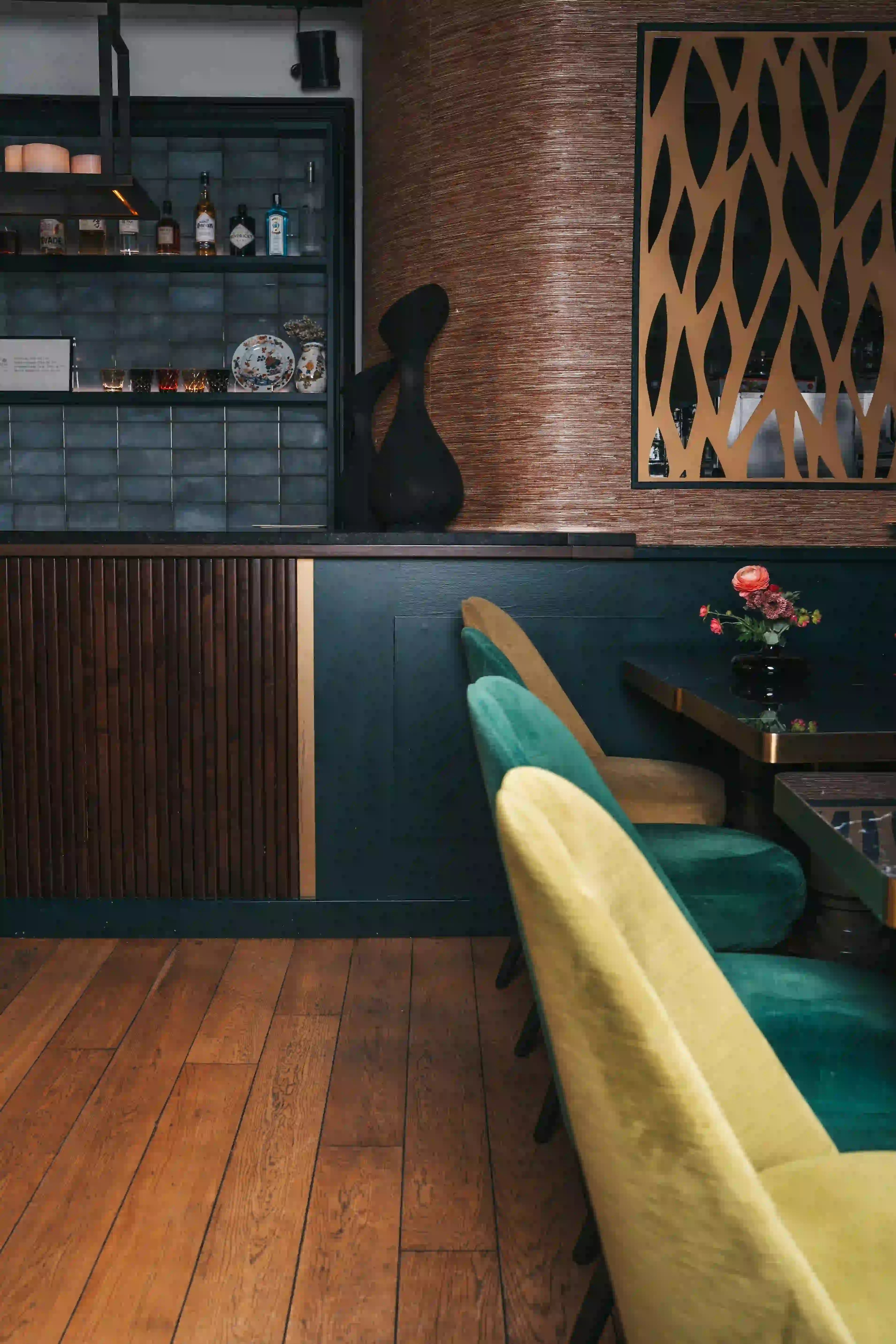 Coin d'un bar avec un sol en bois, des chaises rembourrées de couleur jaune, vert et beige alignées le long d'une table noire, un petit vase avec des fleurs rouges et roses, un mur avec un décor en métal doré et une étagère contenant des bouteilles, verres et objets décoratifs à gauche.