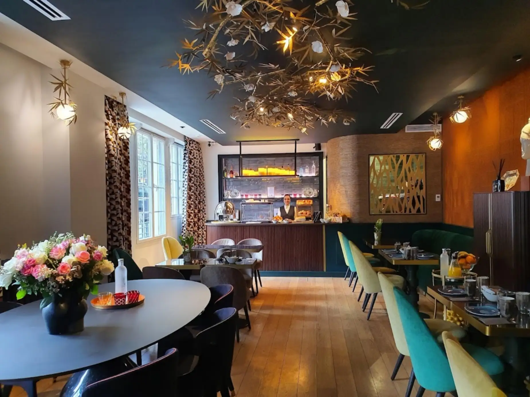 Intérieur élégant d'un café avec grandes fenêtres à gauche, tables noires avec chaises colorées, bouquet de fleurs roses et blanches sur une table ronde devant, et un comptoir en bois au fond où se tient une personne souriante. Les murs sont décorés avec des rideaux à motifs et un miroir décoratif, et un luminaire artistique en forme de branches suspendu au plafond noir illumine la pièce au parquet clair.
