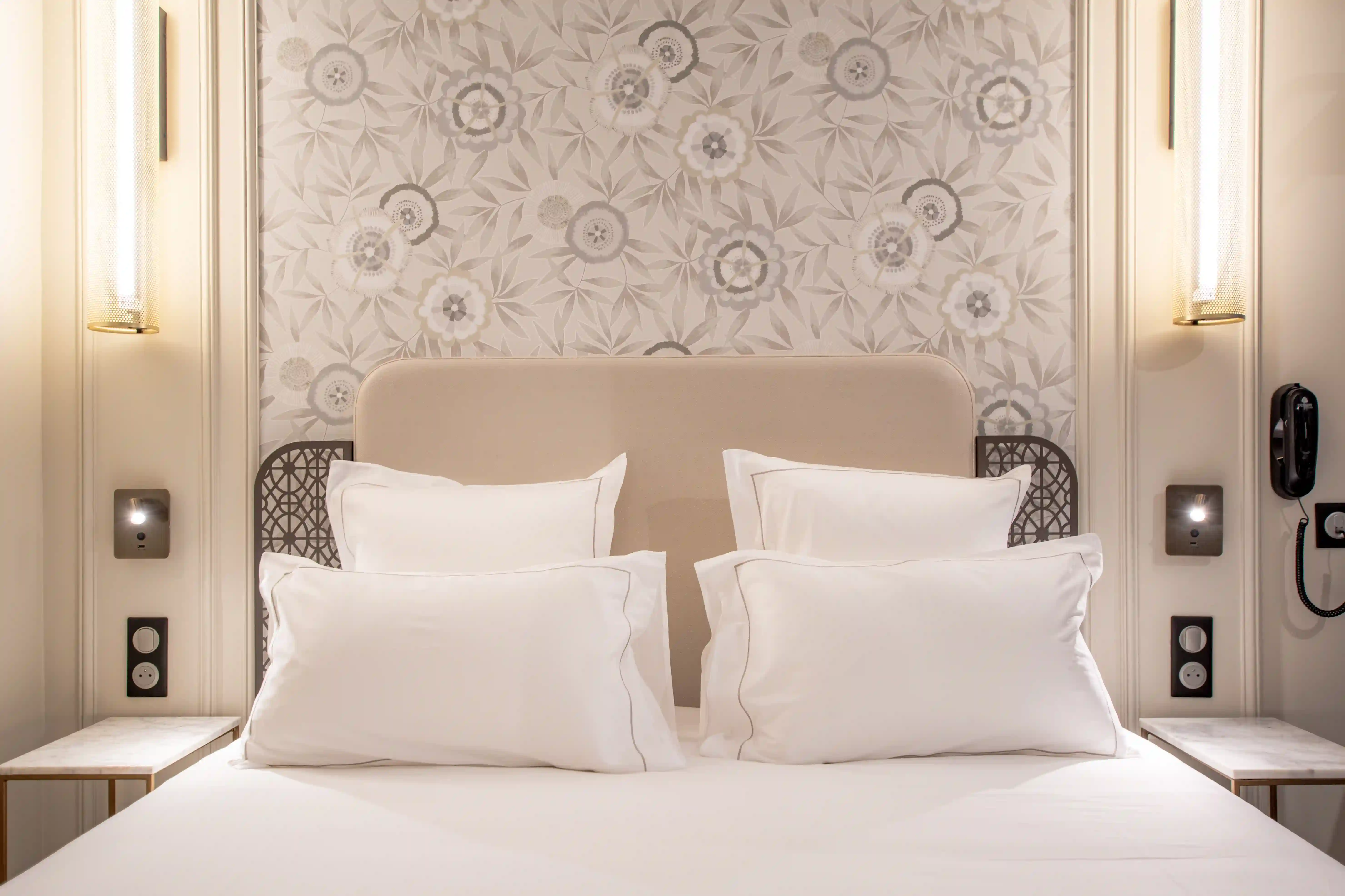 Un lit double avec une tête de lit beige et des coussins blancs disposés en deux rangées, devant un mur avec un papier peint floral gris clair, éclairé par deux appliques murales élégantes et deux tables de chevet en marbre blanc.