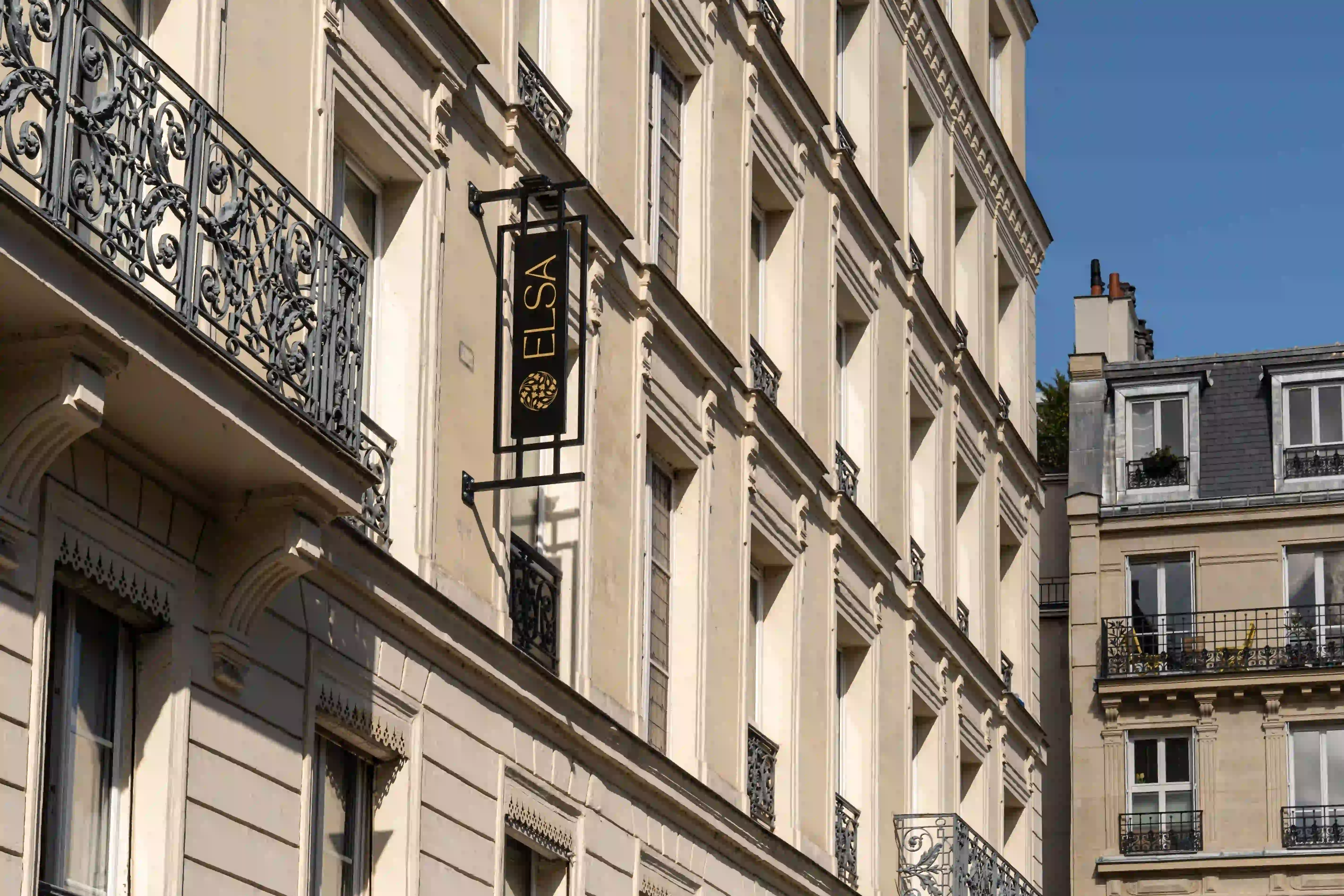 Façades classiques d'immeubles parisiens avec balcons en fer forgé et un panneau noir vertical marqué « ELSA » fixé sur l'un des bâtiments sous un ciel bleu clair.