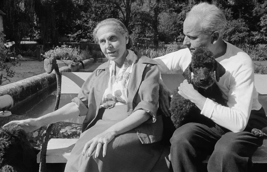 Un couple âgé assis sur un banc extérieur dans un jardin, la femme caressant un chien noir bouclé et l'homme tenant un autre chien similaire sur ses genoux.