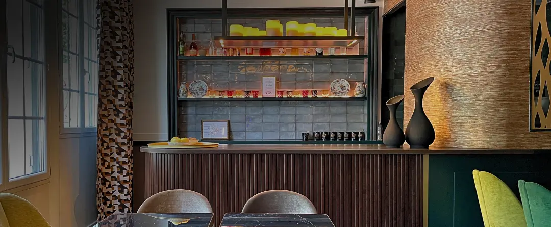 Modern interior space featuring a wooden counter with decorative vases and a shelf behind holding candles, small cups, and decorative plates, with seating including grey and green chairs nearby.