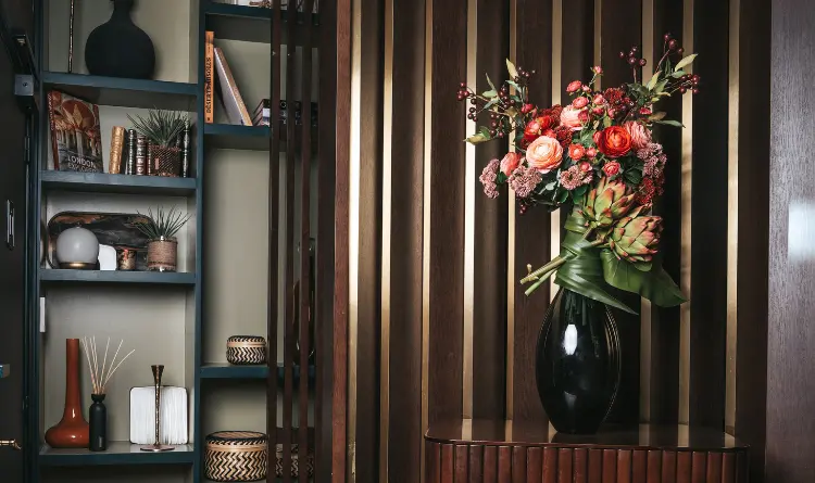 A colorful bouquet of flowers, including roses, red berries and a green artichoke, arranged in a glossy black vase on a wooden surface in front of a dark wood slatted wall