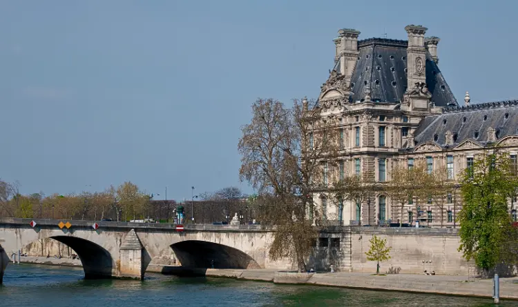 The historic Louvre building on the banks of the Seine with a stone bridge beside it under a clear blue sky in Paris
