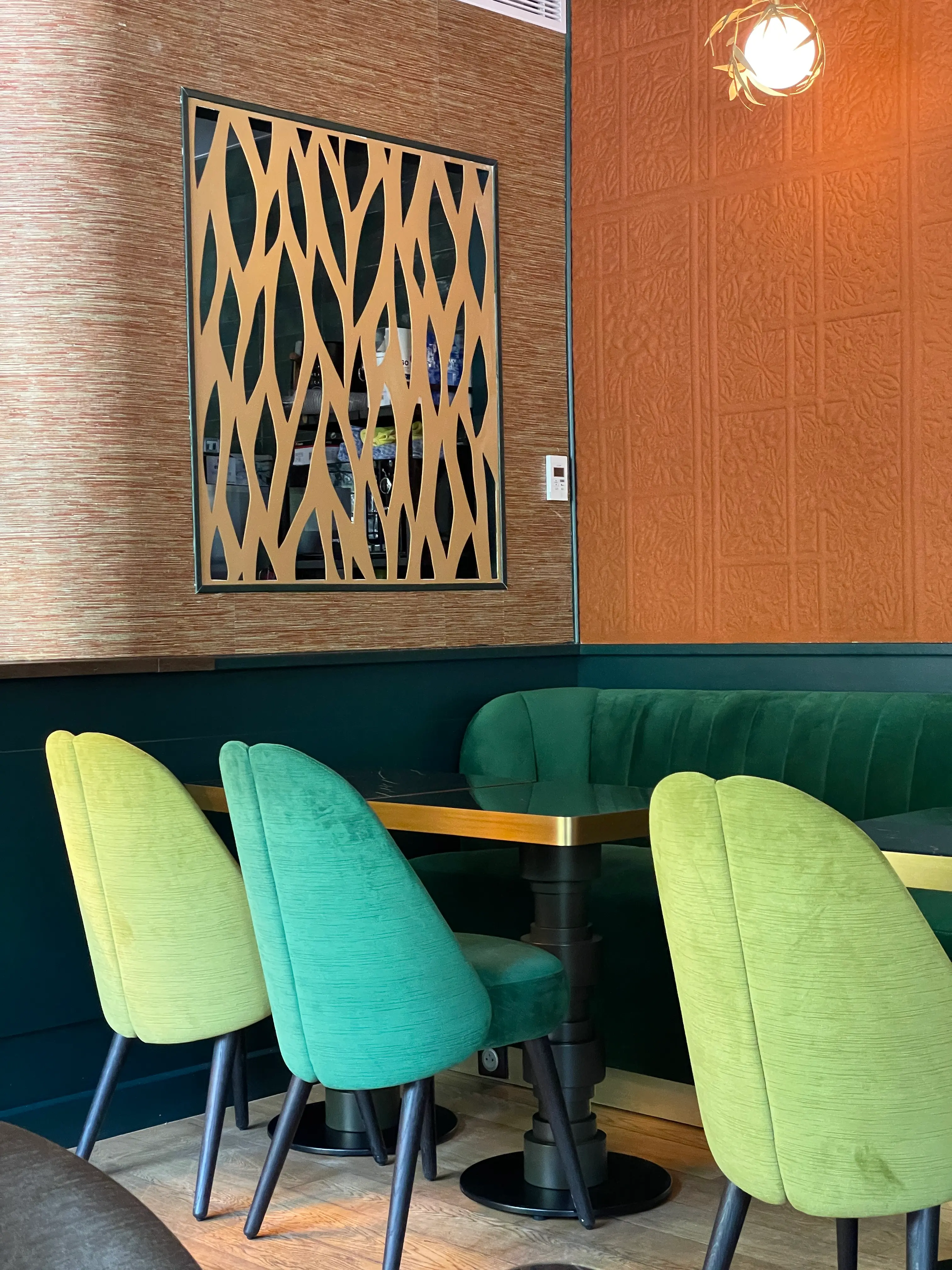 Sitzecke mit einem Tisch, umgeben von hellgrünen und türkisfarbenen Stühlen, einem dunkelgrünen Sofa, einer texturierten orangefarbenen Wand und einer Wanddekoration aus durchbrochenem Holz