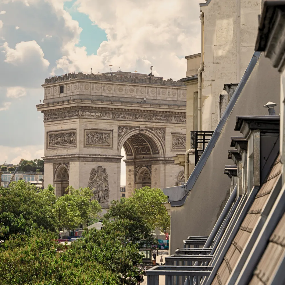 Vista parcial do Arco do Triunfo em Paris rodeado por árvores verdes, com edifícios residenciais cinzentos e beges do lado direito sob um céu nublado.