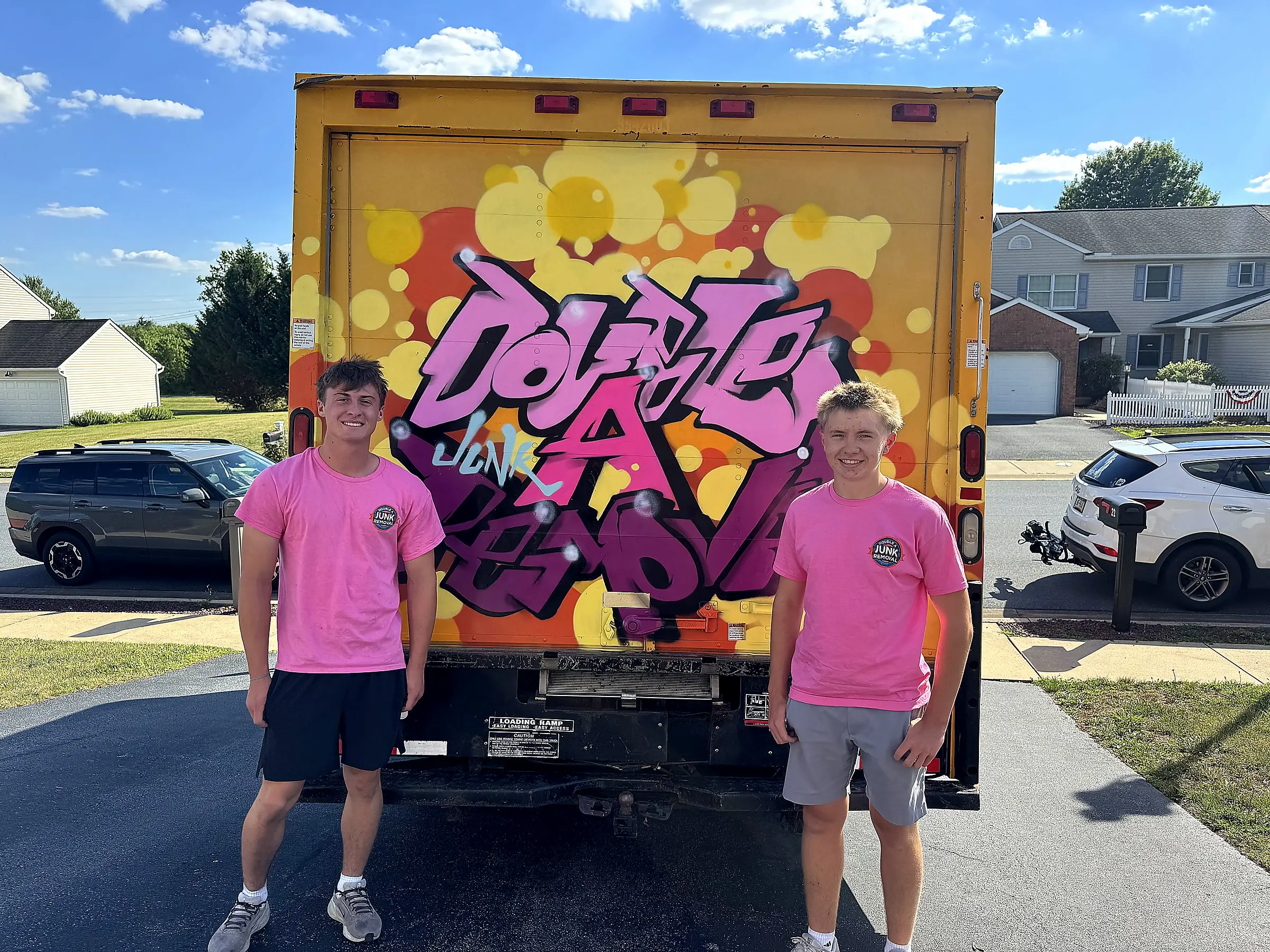 Two young men in pink shirts with a junk removal logo stand smiling in front of a yellow truck with graffiti-style colorful text and patterns on the back door in a residential neighborhood on a sunny day.