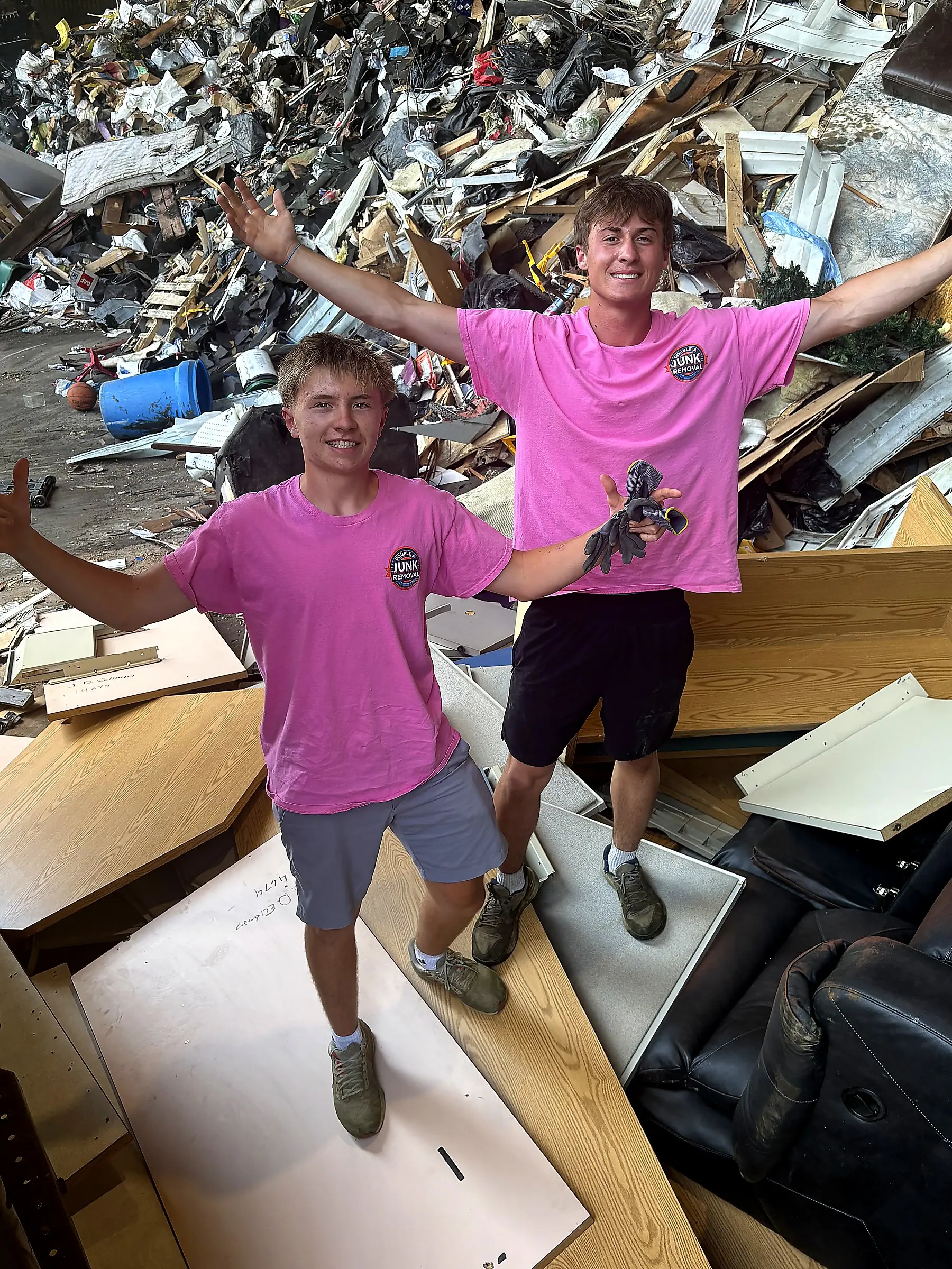Two young men wearing matching pink T-shirts with 'Junk Removal' logo pose with arms spread wide amidst a large pile of scattered furniture and trash indoors.