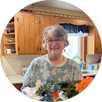 Smiling elderly woman with glasses standing in a kitchen holding a plant with colorful flowers.